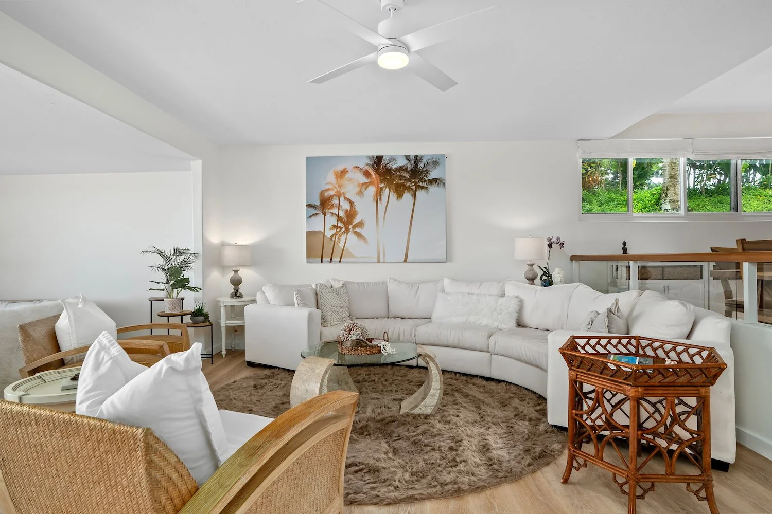 Bright living room at Mahina Kai Kauai with white sectional sofa, glass coffee table, rattan side table, tropical decor, and ocean sunset wall art, with large window showing greenery outside.