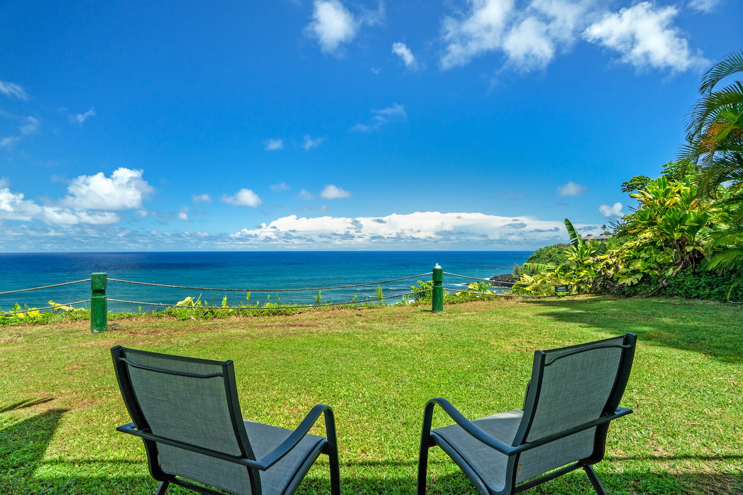 Two chairs facing a lush green lawn with a view of the ocean, blue sky with clouds, and tropical foliage in the background.
