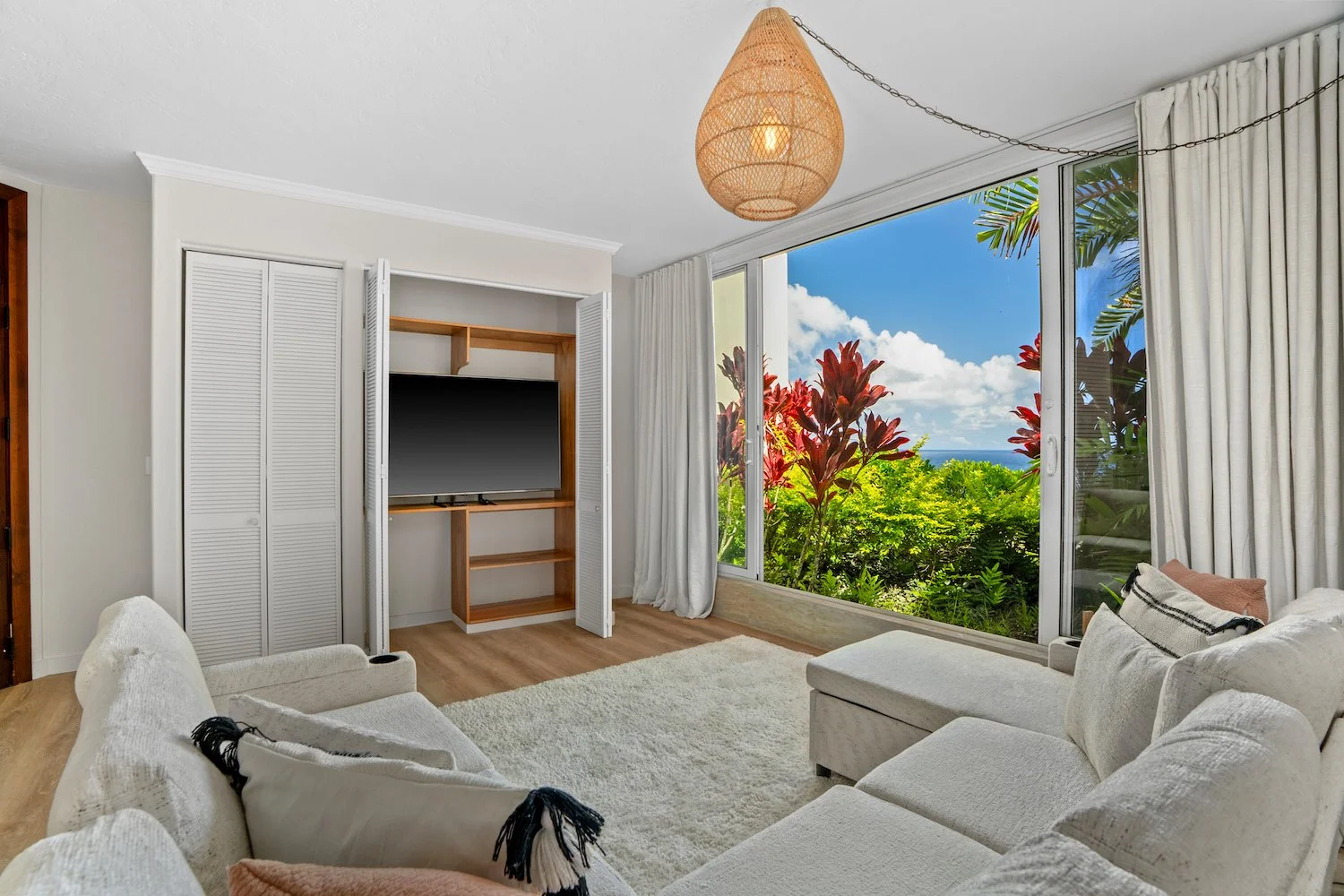Living room with a large window overlooking greenery and blue sky, white couches, a television in a wooden nook, white louvered closet doors, and a woven pendant light.