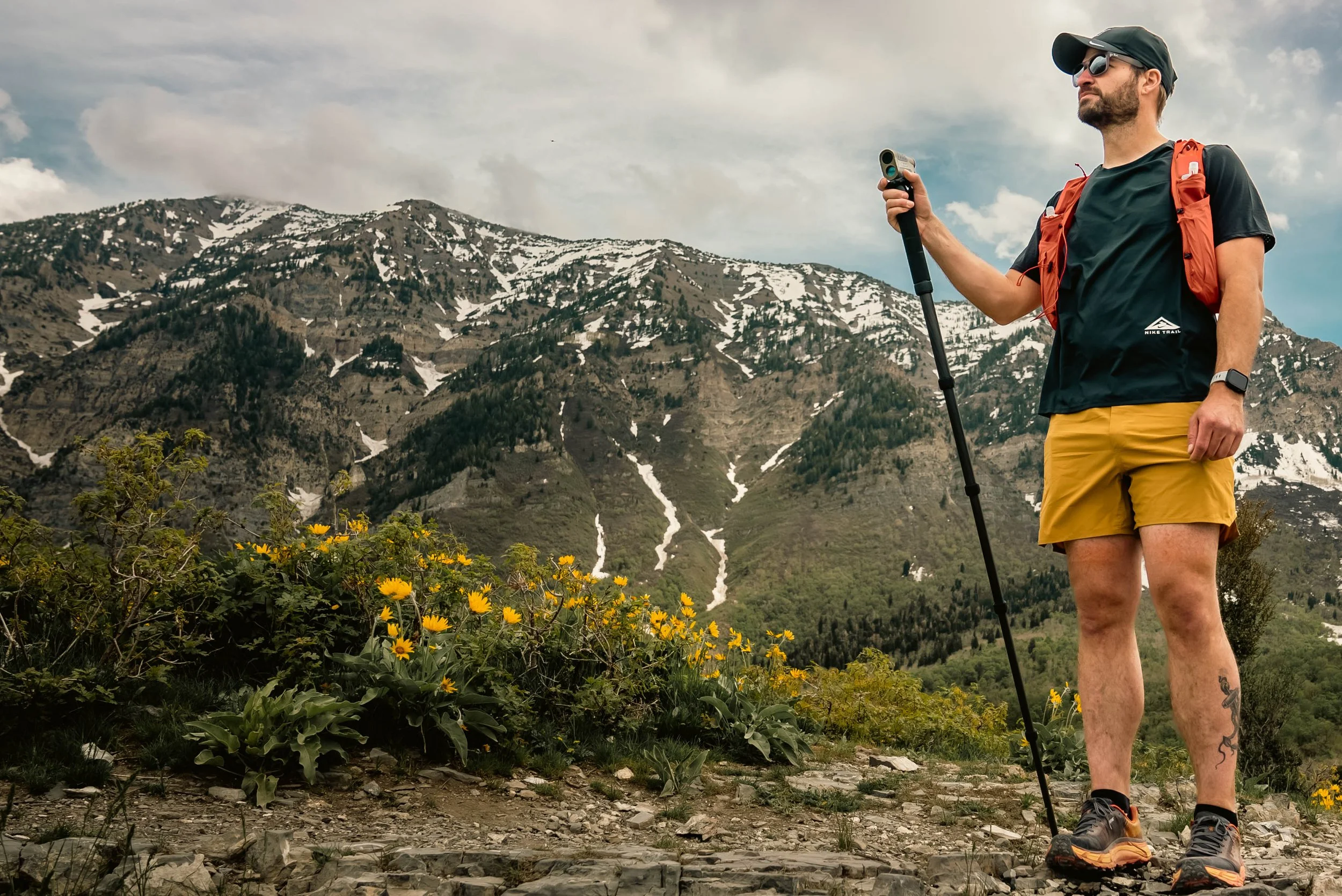 A male runner holding a monopod and range finder with yellow flowers with the snowcapped Wasatch mountain range behind him.