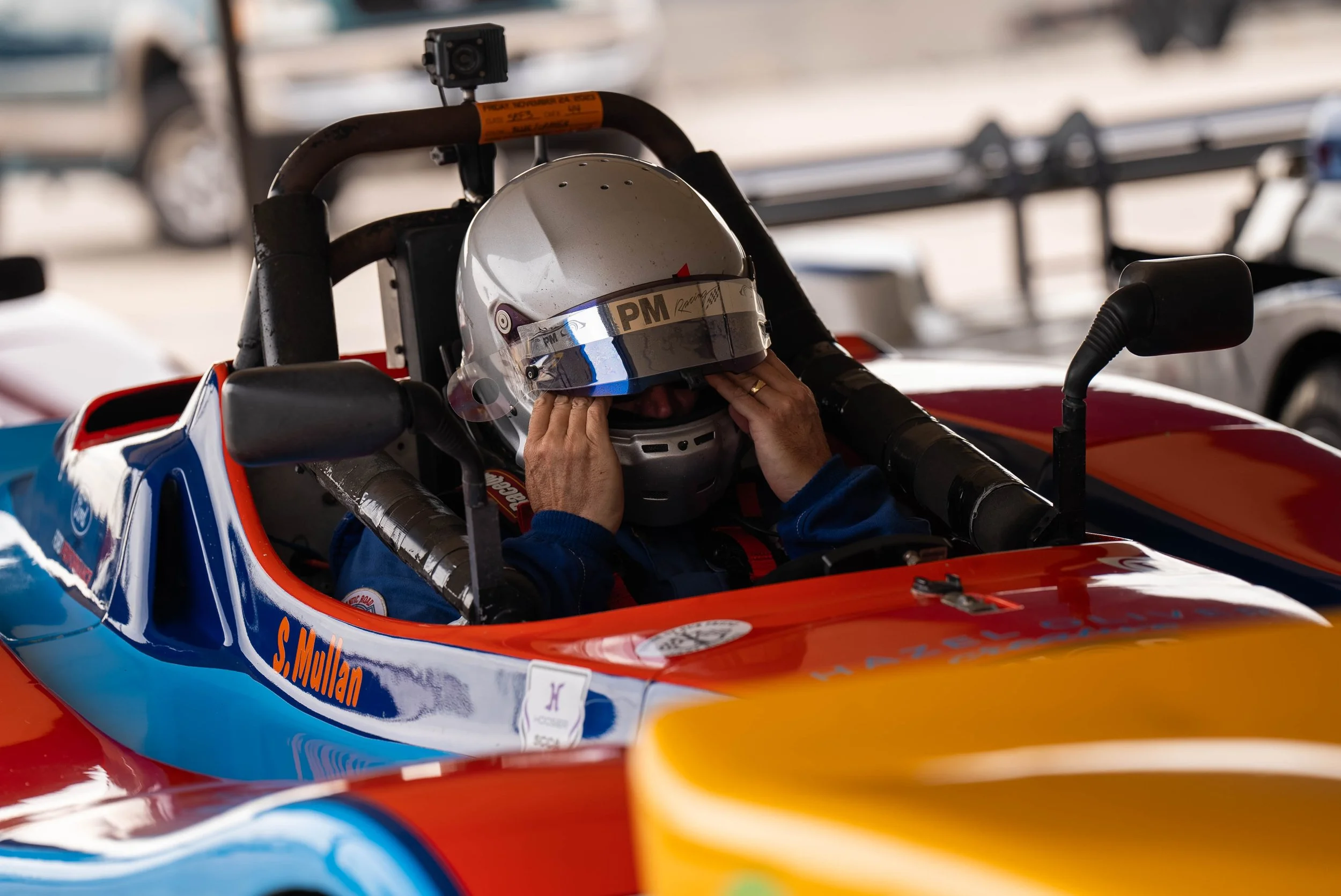 A racer driver sitting in his orange and blue Spec Racer Ford adjusting his helmet at an SCAA race in Southern Florida.