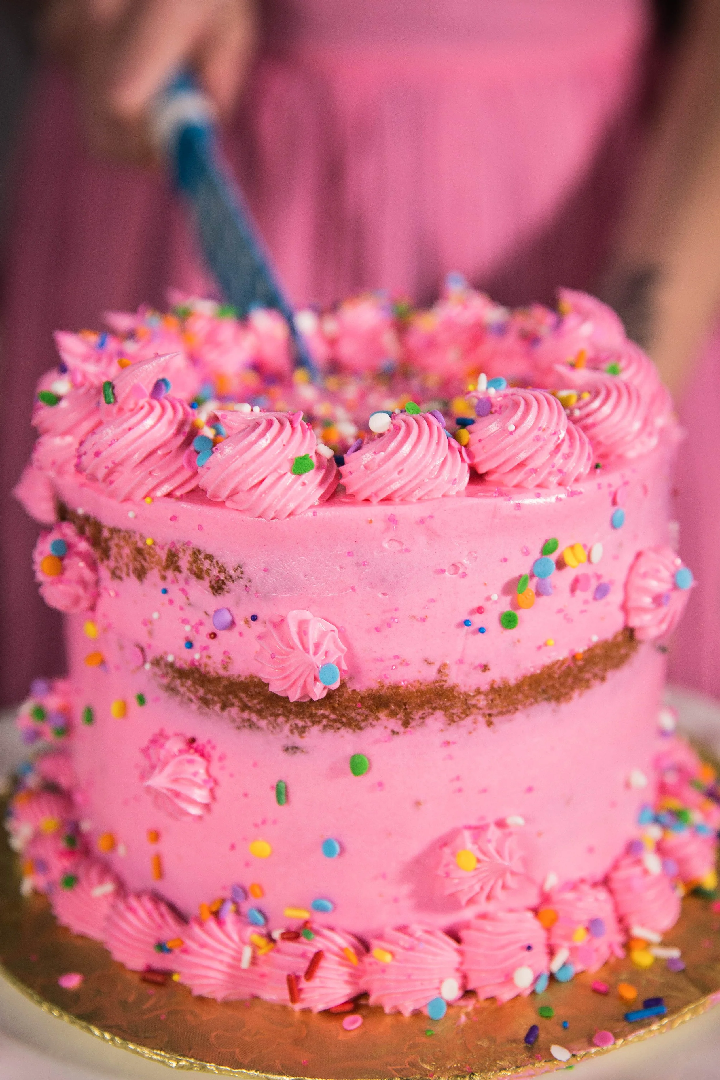 A bright pink iced cake with rainbow sprinkles getting cut into with a blue knife at a local bakery.