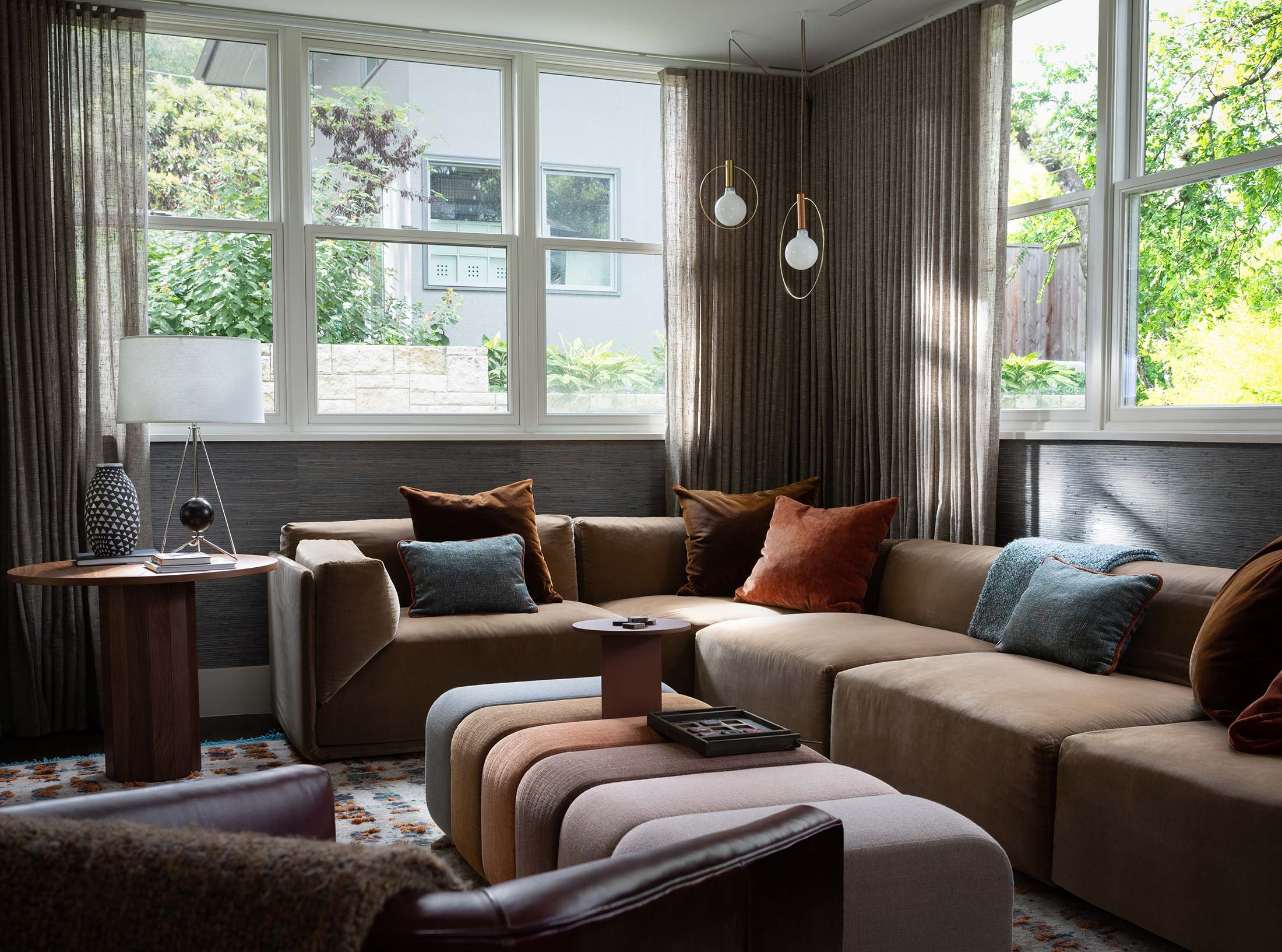 The photograph showcases a cozy lounge area bathed in natural light from multiple windows. A sectional sofa in a soft taupe tone offers ample seating, adorned with throw pillows in rich brown, burnt orange, and teal for a pop of color. In the corner 