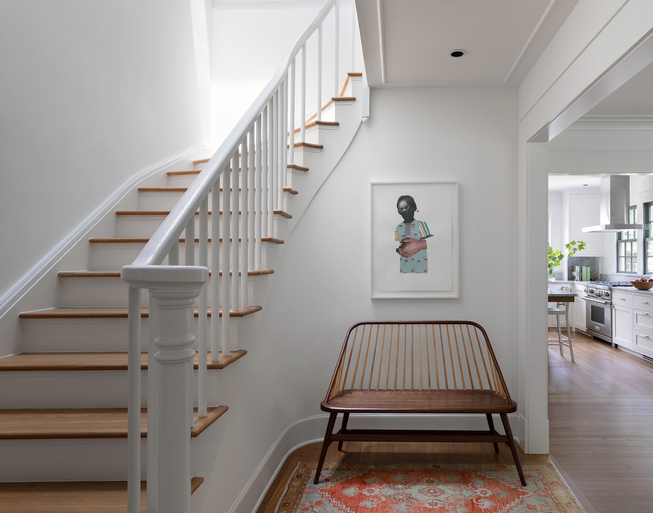 This hallway exudes a minimalist elegance, punctuated by the presence of a mid-century modern bench that anchors the space. The bench features a slender wooden frame with a rich, dark stain, which supports a series of vertical slats forming the backr