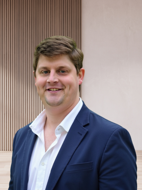 A smiling man in a navy blazer and white shirt standing outdoors in front of a modern building with wooden and concrete panels.