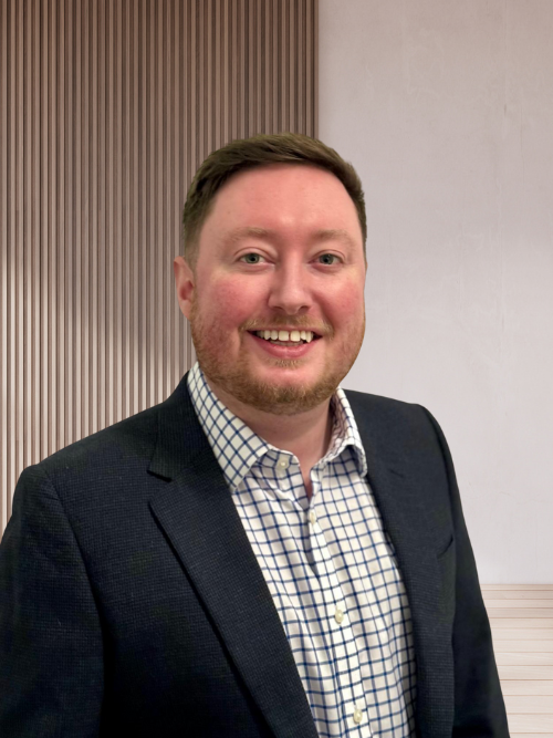 A smiling man in a blazer and checkered shirt standing indoors against a background with vertical wood panels and a plain wall.