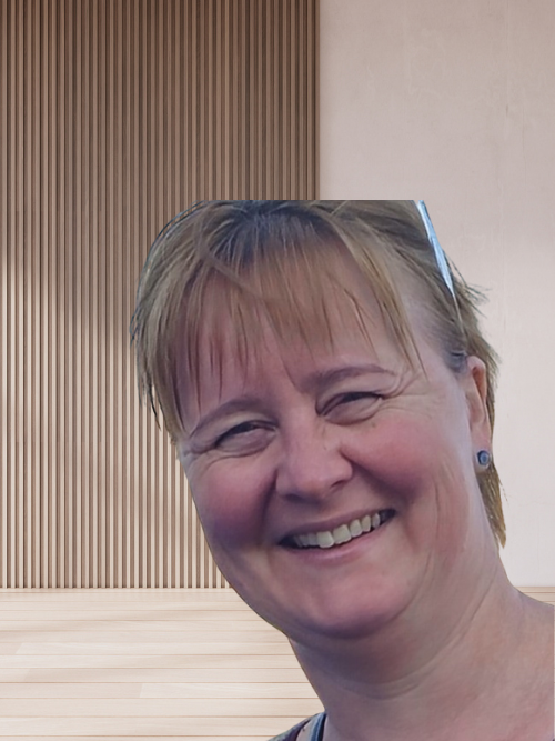 A woman with short blonde hair smiling outdoors, with a wooden and beige wall in the background.