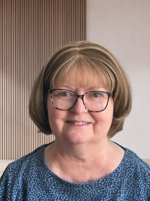 A middle-aged woman with light brown hair and glasses smiling at the camera, wearing a blue patterned top, with a wooden and light-colored wall background.