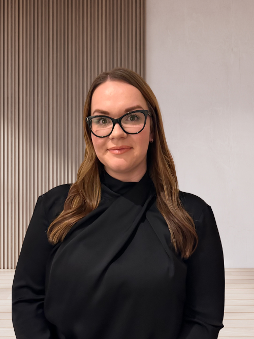 A woman with long brown hair, wearing black glasses and a black top, standing indoors in front of a background with wooden slats and a plain wall.