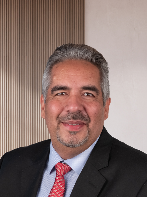 A middle-aged man with gray hair, a mustache, and beard, wearing a black suit, light blue shirt, and red patterned tie, smiling at the camera against a background with a vertical wood panel and a cream-colored wall.