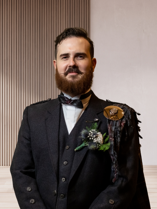 A man with a beard and mustache wearing a vintage-style black suit, bow tie, and a boutonniere, standing indoors against a background with vertical wood paneling and plain wall.