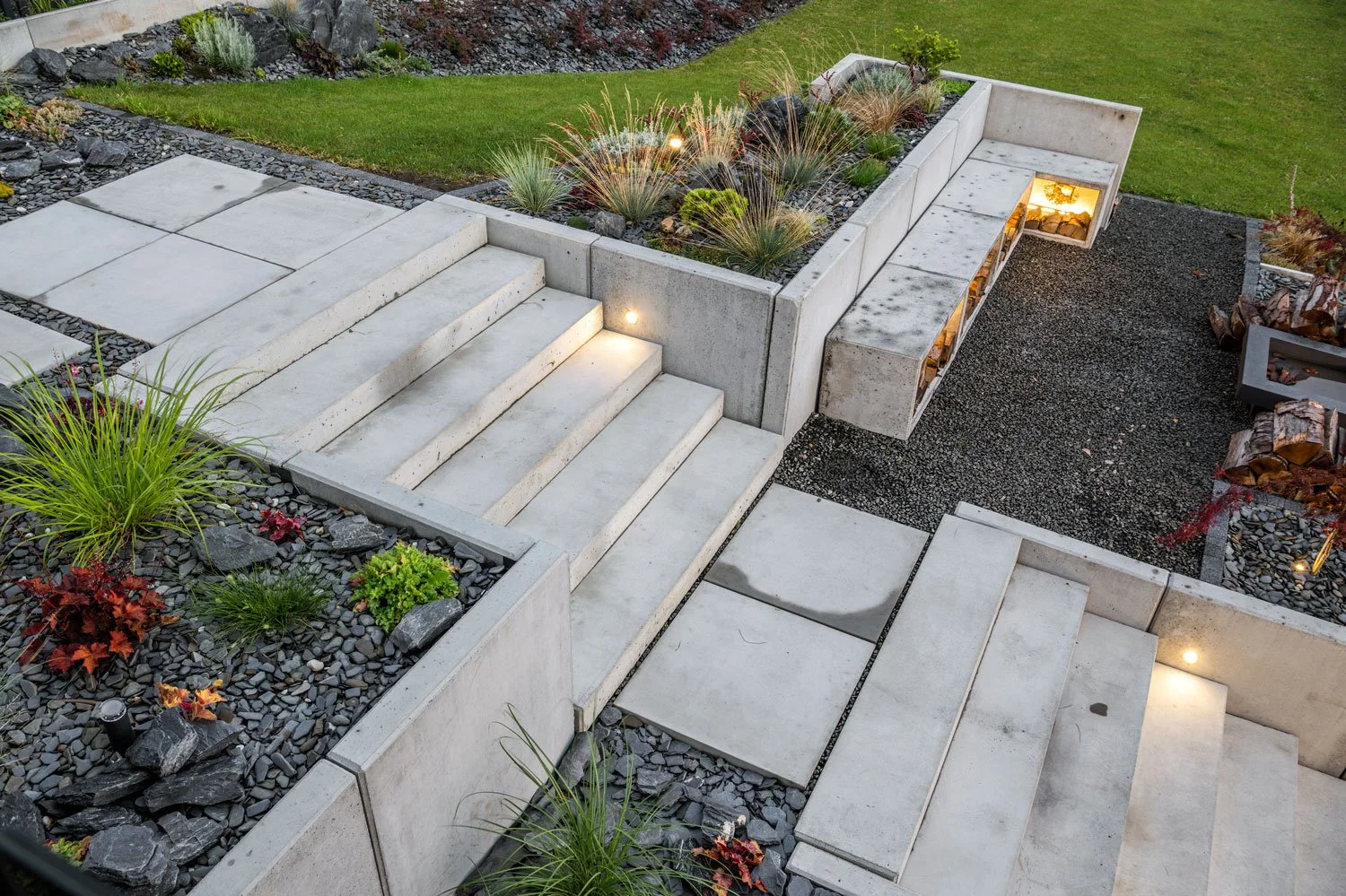 Modern outdoor staircase with concrete steps and built-in lighting, surrounded by landscaped garden beds with rocks, ornamental grasses, and small plants.