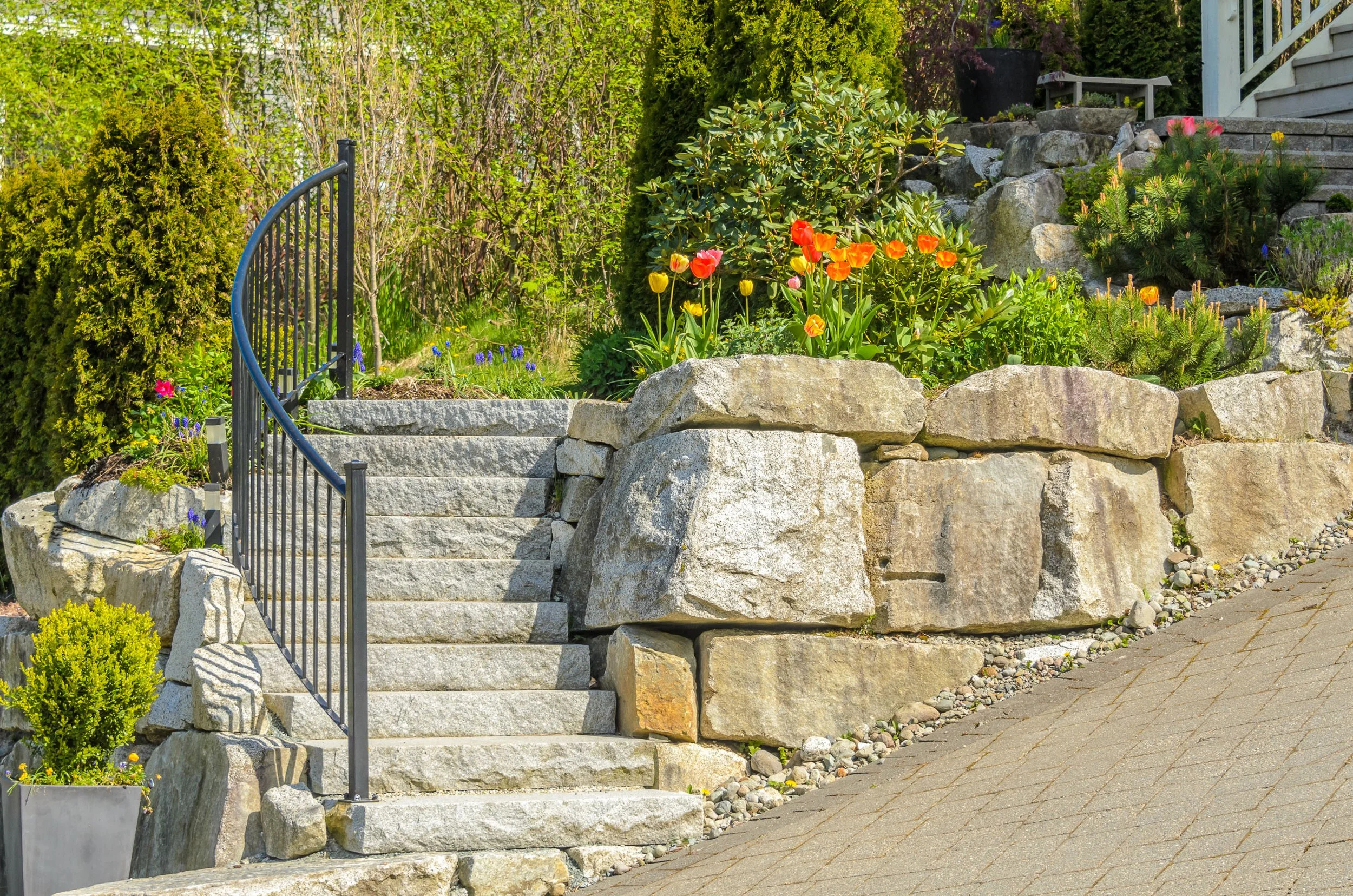 Stone staircase leading to a garden with various plants and shrubs, including orange and yellow flowers, surrounded by rocks and a black metal railing on the left side.