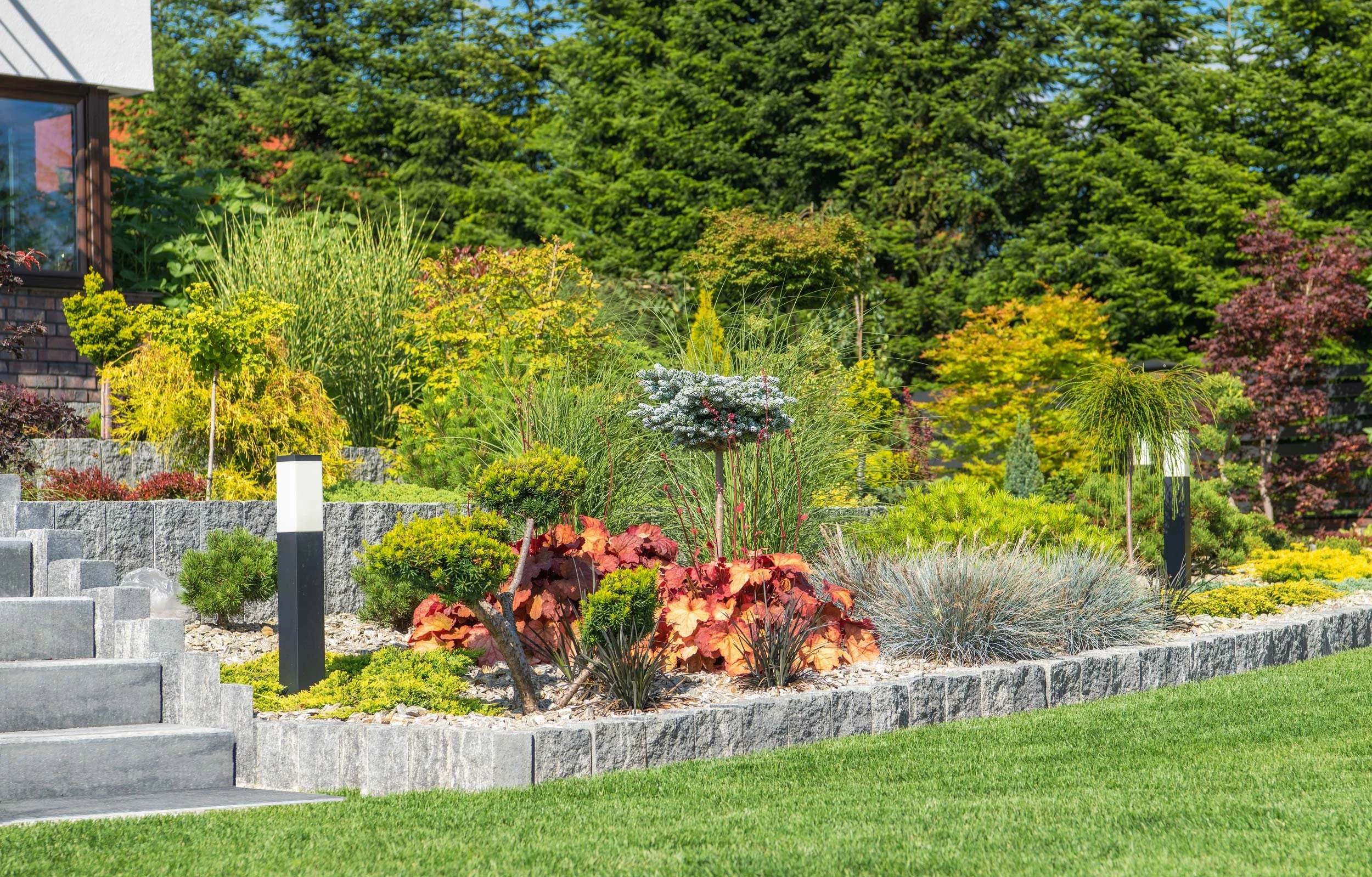 A landscaped garden area with a variety of plants and shrubs, a stone retaining wall, and lush green trees in the background.
