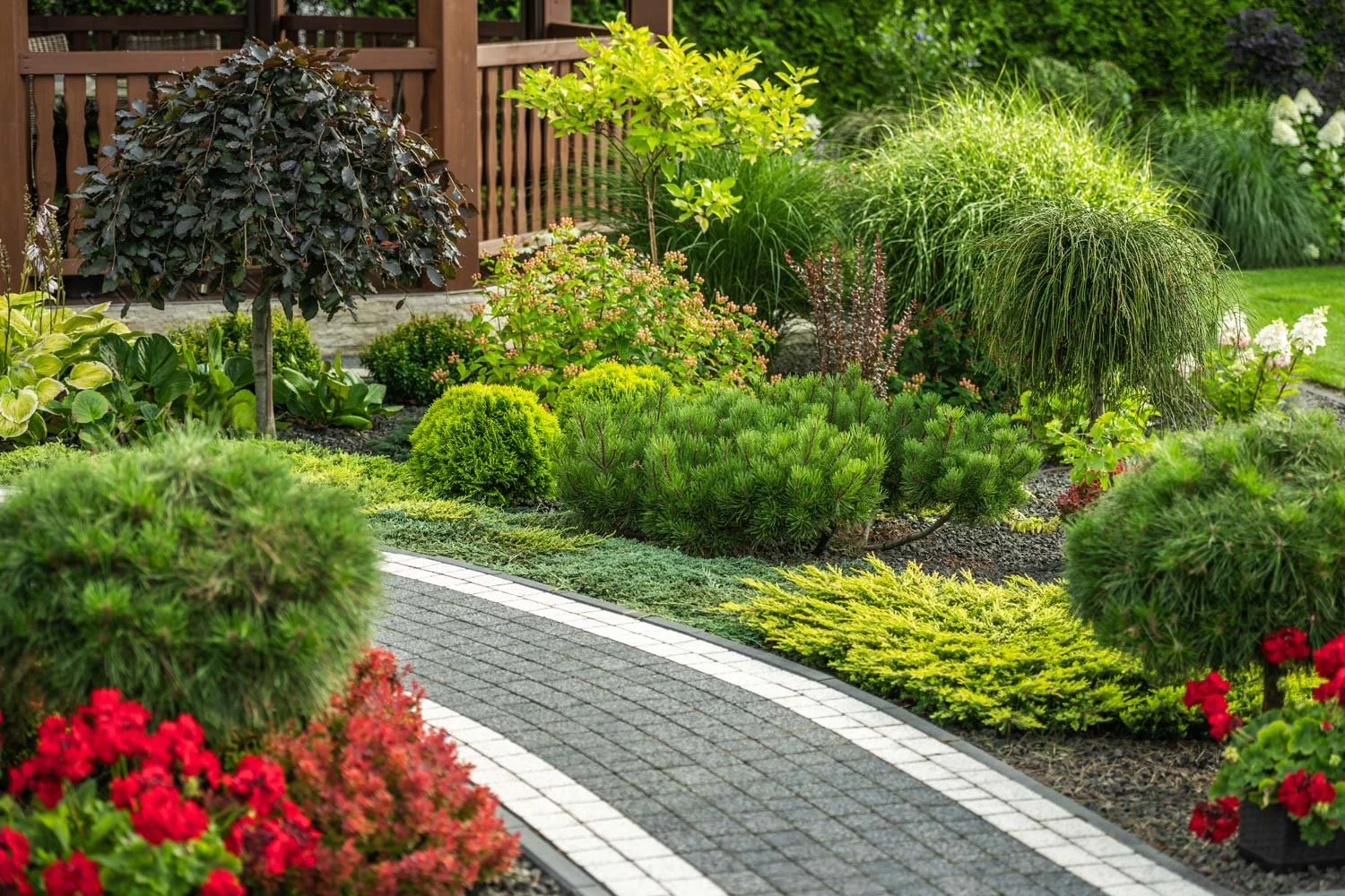 A well-maintained garden with various shrubs, bushes, and flowering plants along a curved pathway with gray bricks and white borders, enclosed by a wooden fence.