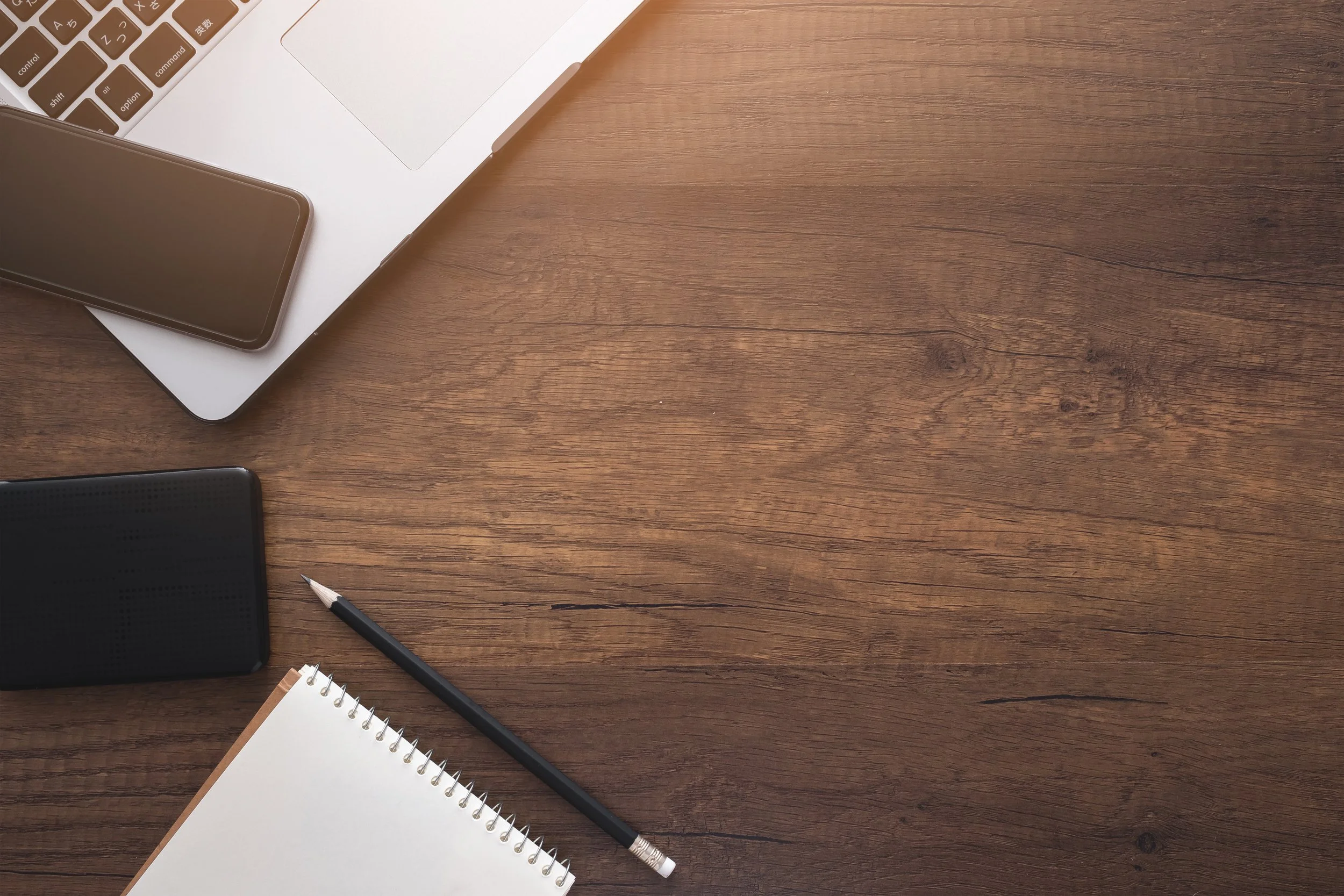 A wooden desk with a silver laptop, a black smartphone, a black portable speaker, a spiral notepad, and a black pen.