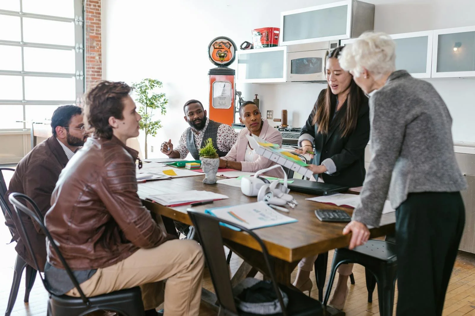 A diverse group of six people in a meeting room, engaging in a discussion. One woman is presenting and holding a document or chart, while others are listening at a large wooden table with notebooks, papers, and a potted plant. The room has large windows, white cabinetry, and a brick wall in the background.