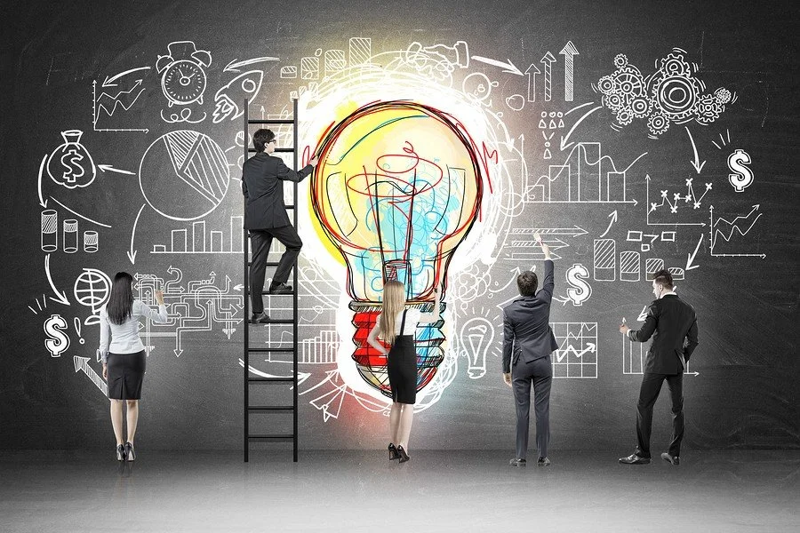 Business professionals drawing and working on a large illuminated light bulb and various business and growth charts on a blackboard.