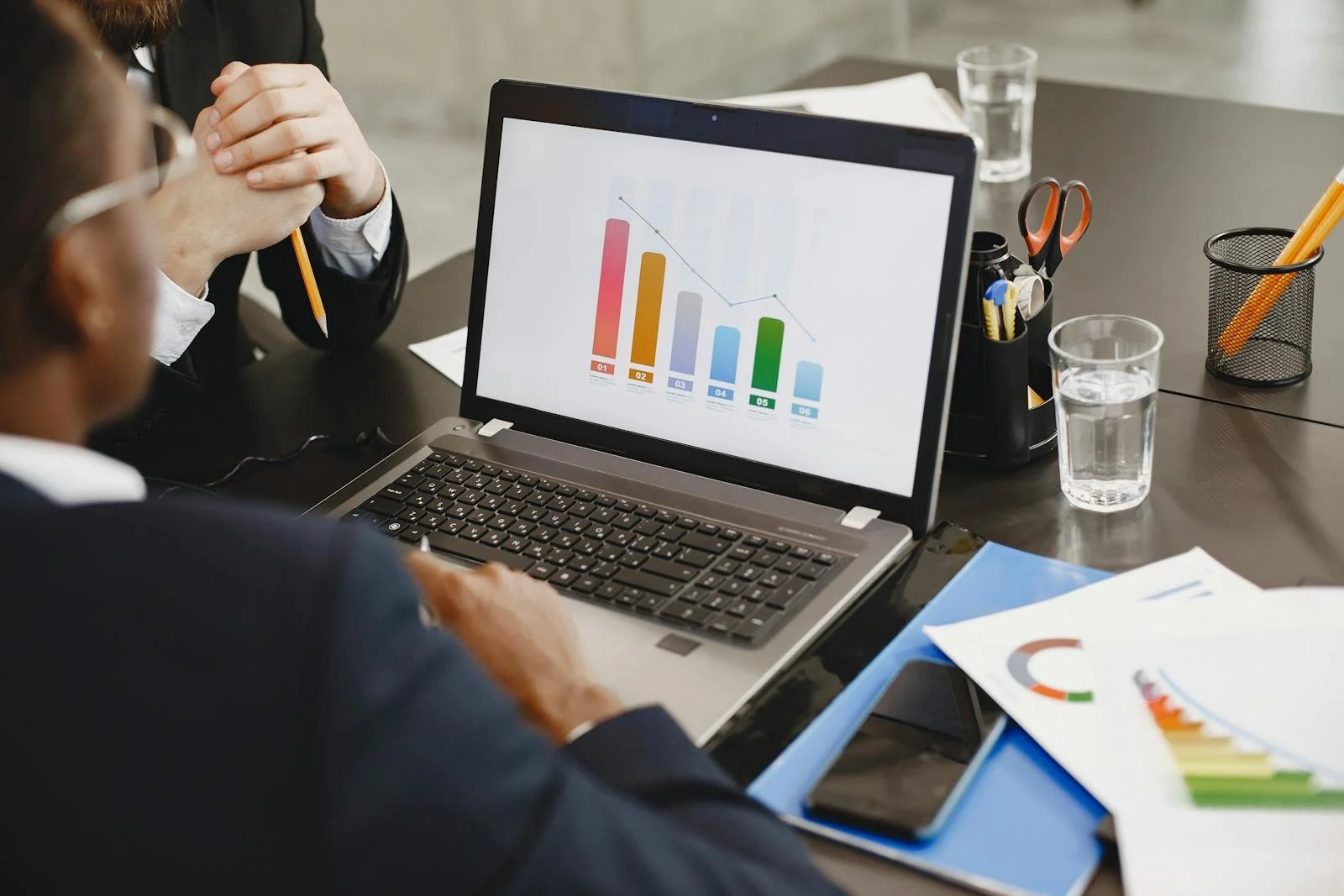 People working on a business project with graphs and charts on tools like a laptop, papers, and a smartphone, with glasses of water on the table.