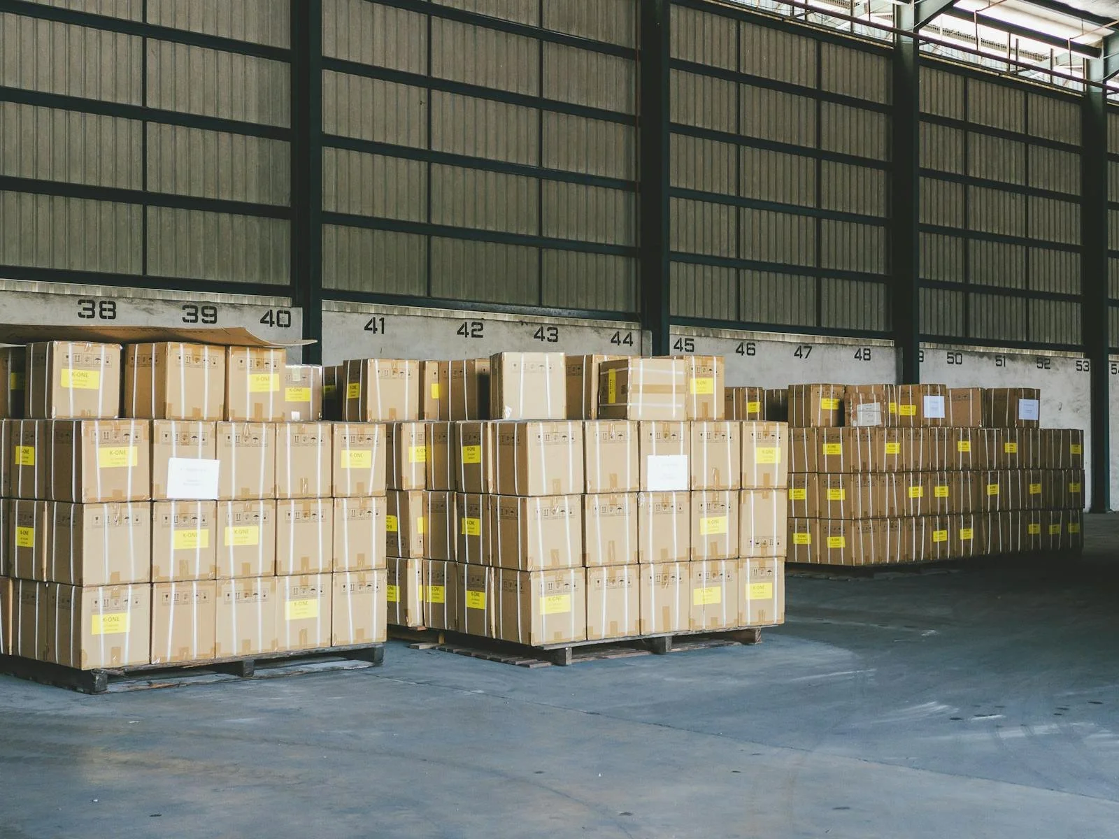 Warehouse with stacks of cardboard boxes on pallets, labeled with yellow tags, arranged in rows, and a wall with numbered sections in the background.