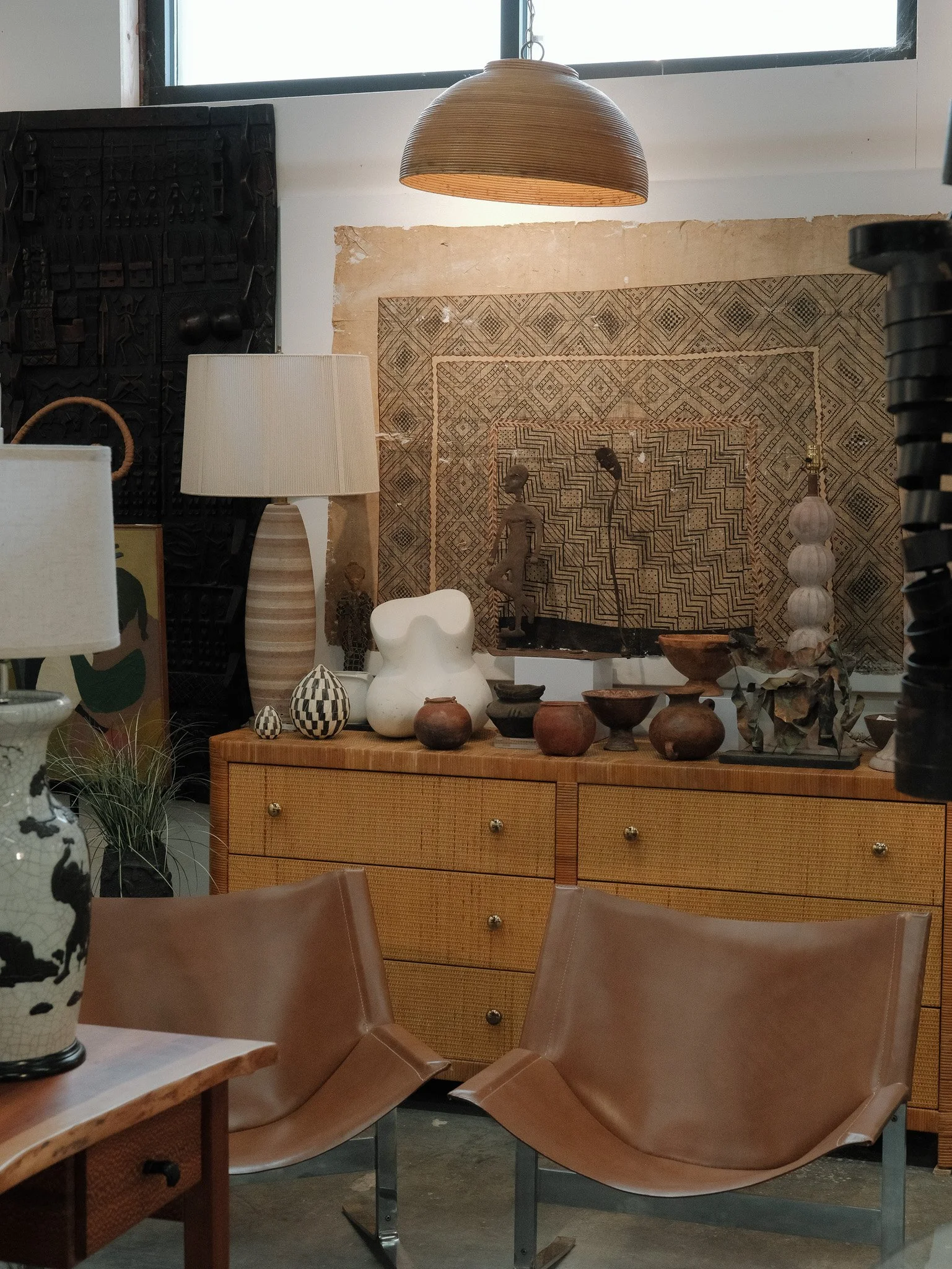 Interior of a room with a mid-century modern wooden dresser, various decorative pottery and sculptures, a large textured wall hanging, two tan leather chairs, a table lamp, a floor lamp, and a large window above.