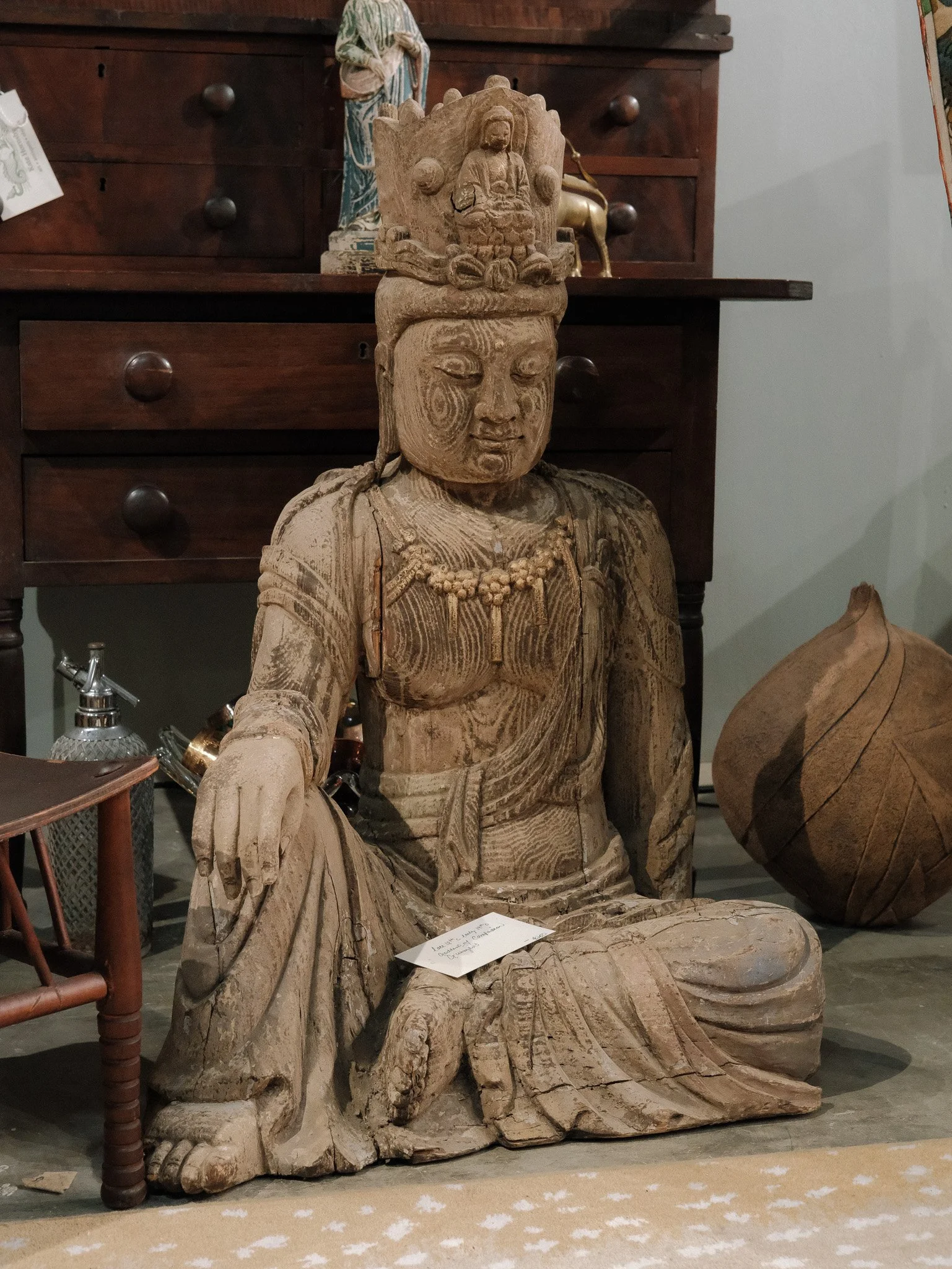 Wooden sculpture of a woman in traditional attire, sitting with one leg folded and the other extended, with a smaller figure on her head.