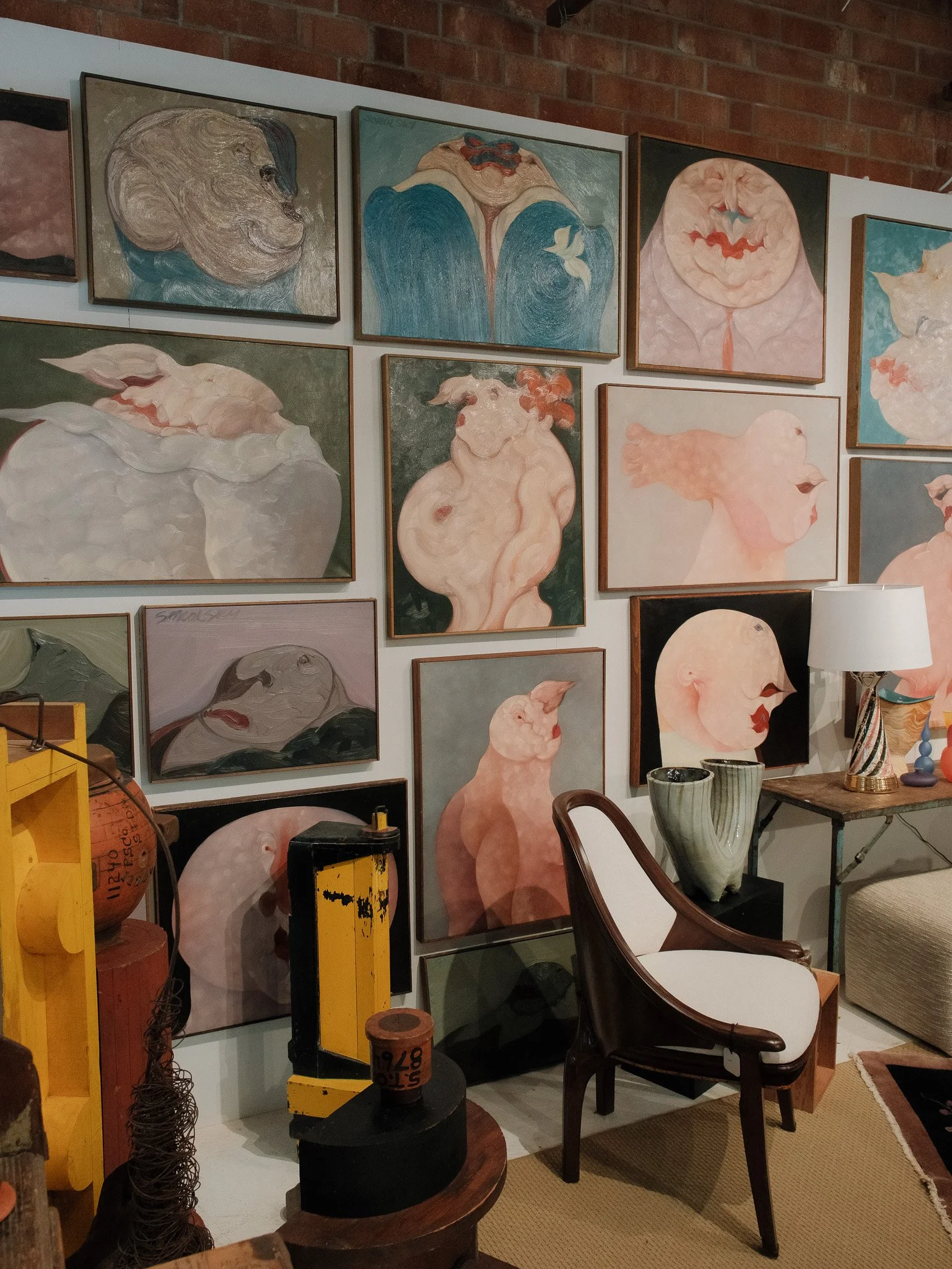 Gallery wall featuring multiple abstract paintings of humanoid figures with exaggerated features and pastel color palette, accompanied by eclectic furniture and decorative objects.