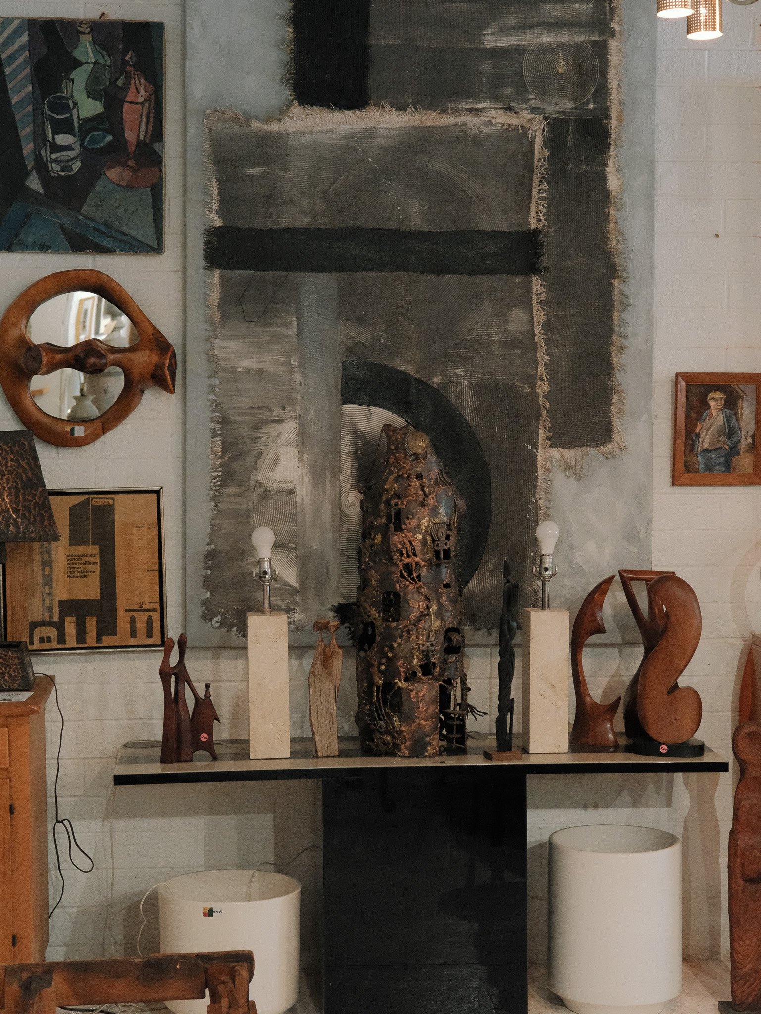 A display of various sculptures and artworks in a gallery, featuring a large abstract painting in the background and sculptures made of wood, metal, and stone on a black shelf.