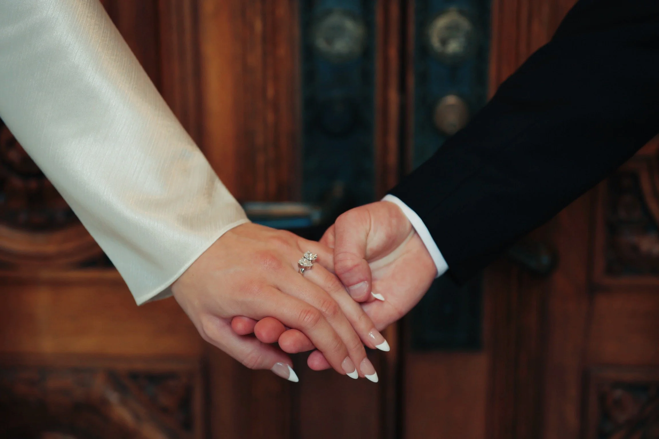 Close-up of a newlywed couple holding hands, showing the bride's engagement and wedding rings, with a wooden background.