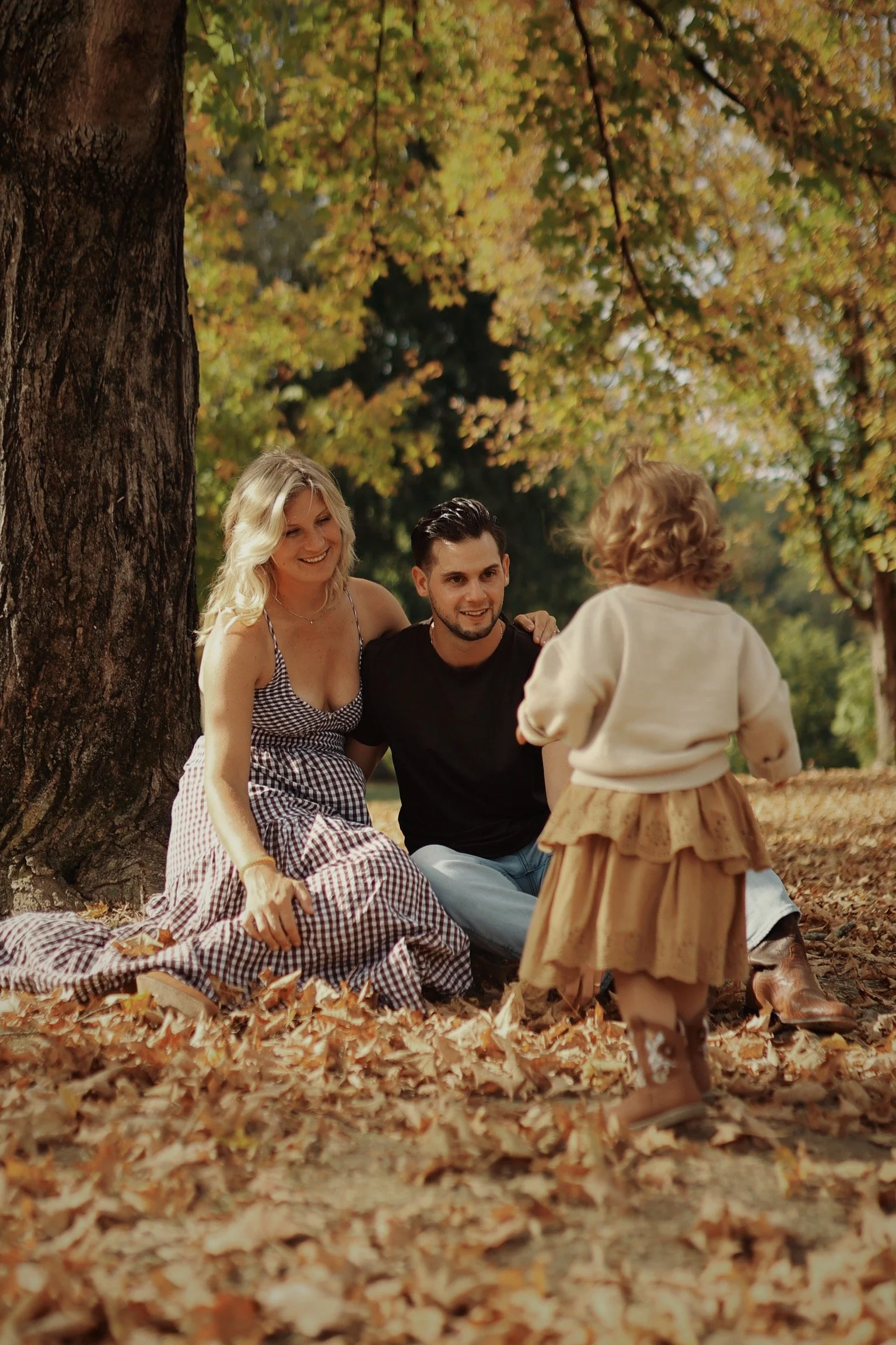 A family of three sitting on autumn leaves beneath a large tree, enjoying the outdoors. The woman and man are smiling at a young girl standing in front of them, surrounded by fall foliage.