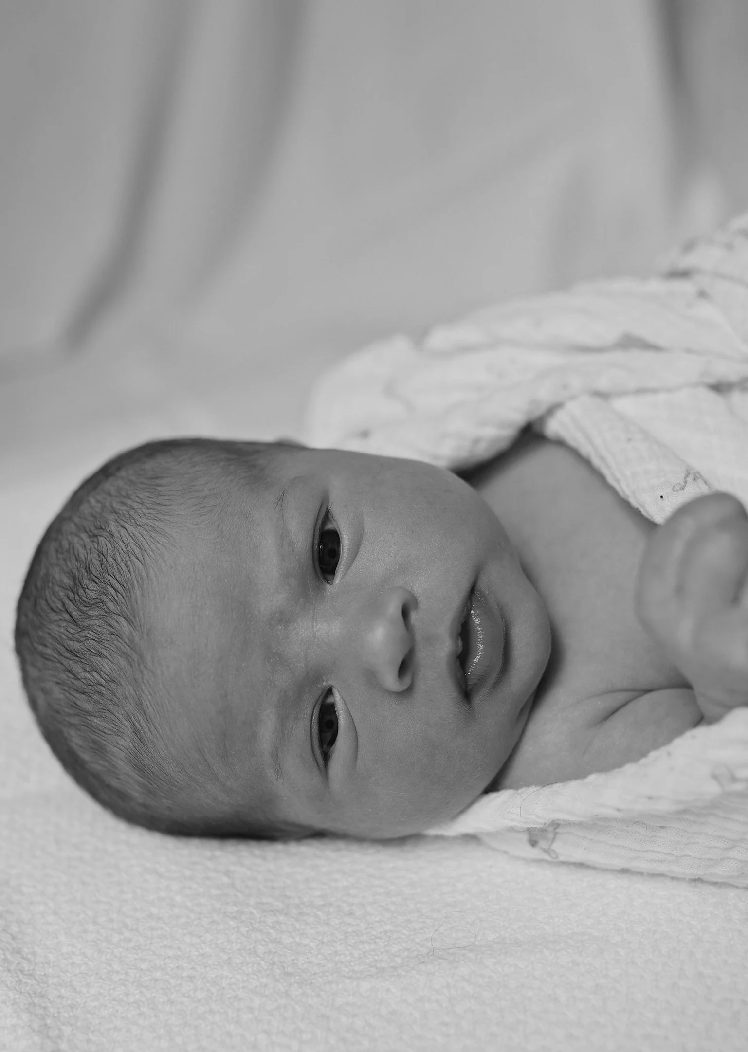 Black-and-white photo of a newborn baby lying on a soft surface, looking towards the camera with wide eyes and a curious expression.