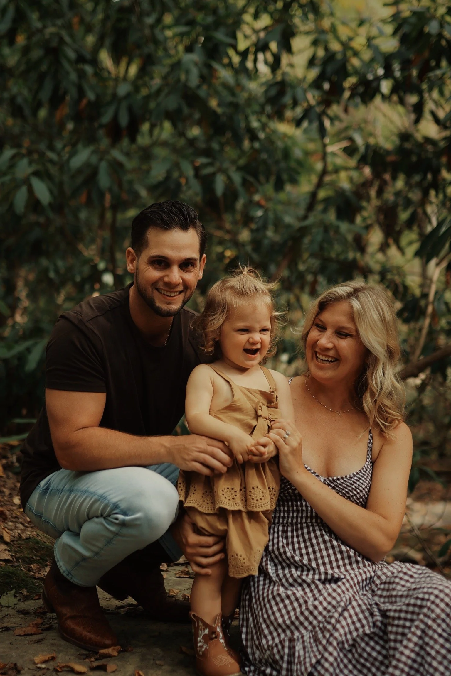 Family of three smiling and laughing outdoors in a forest, with a man crouching, a woman sitting, and a young girl standing between them.