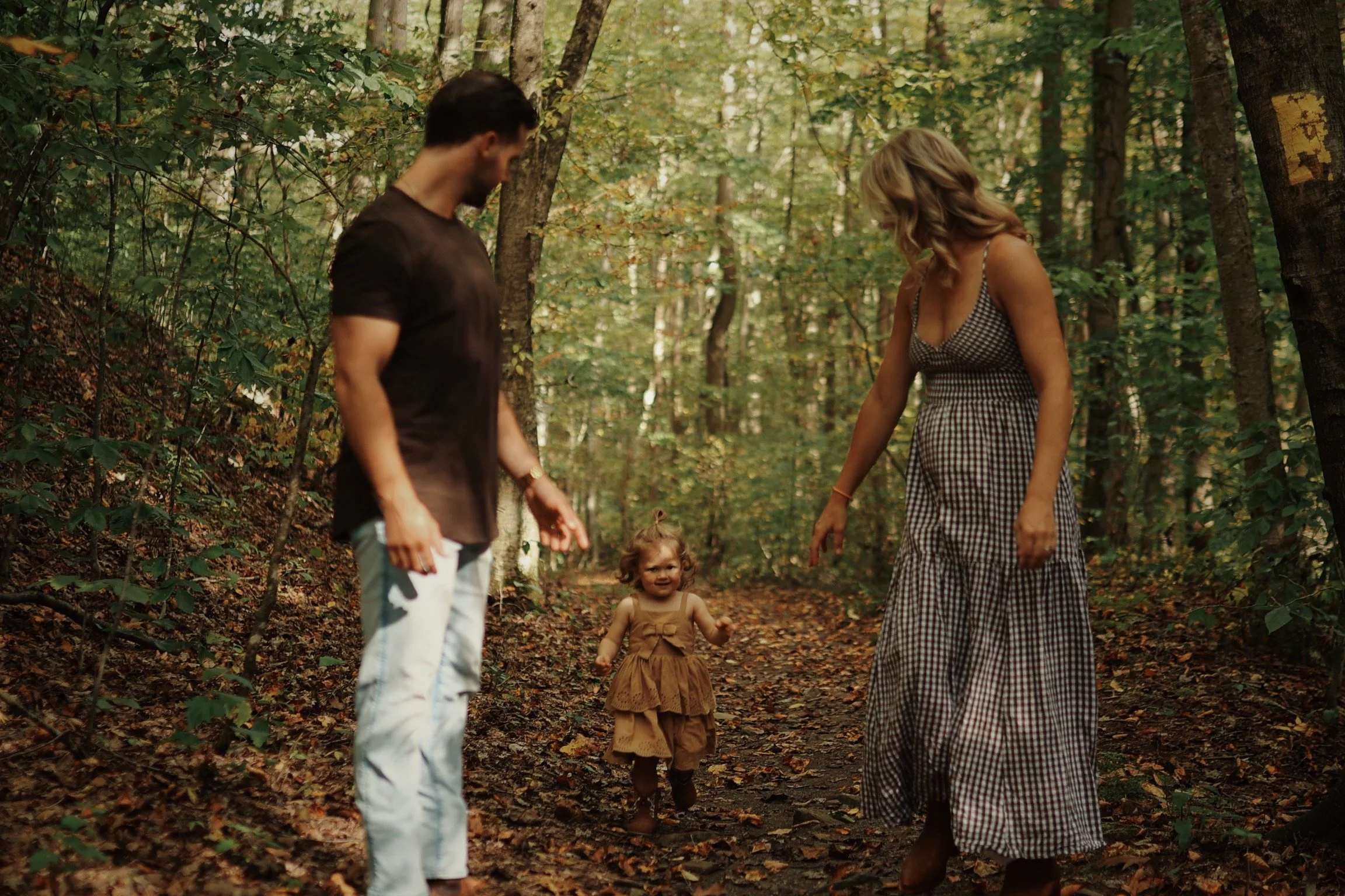 A family of three walking on a forest trail during fall, with a toddler girl in the middle and her parents on either side.