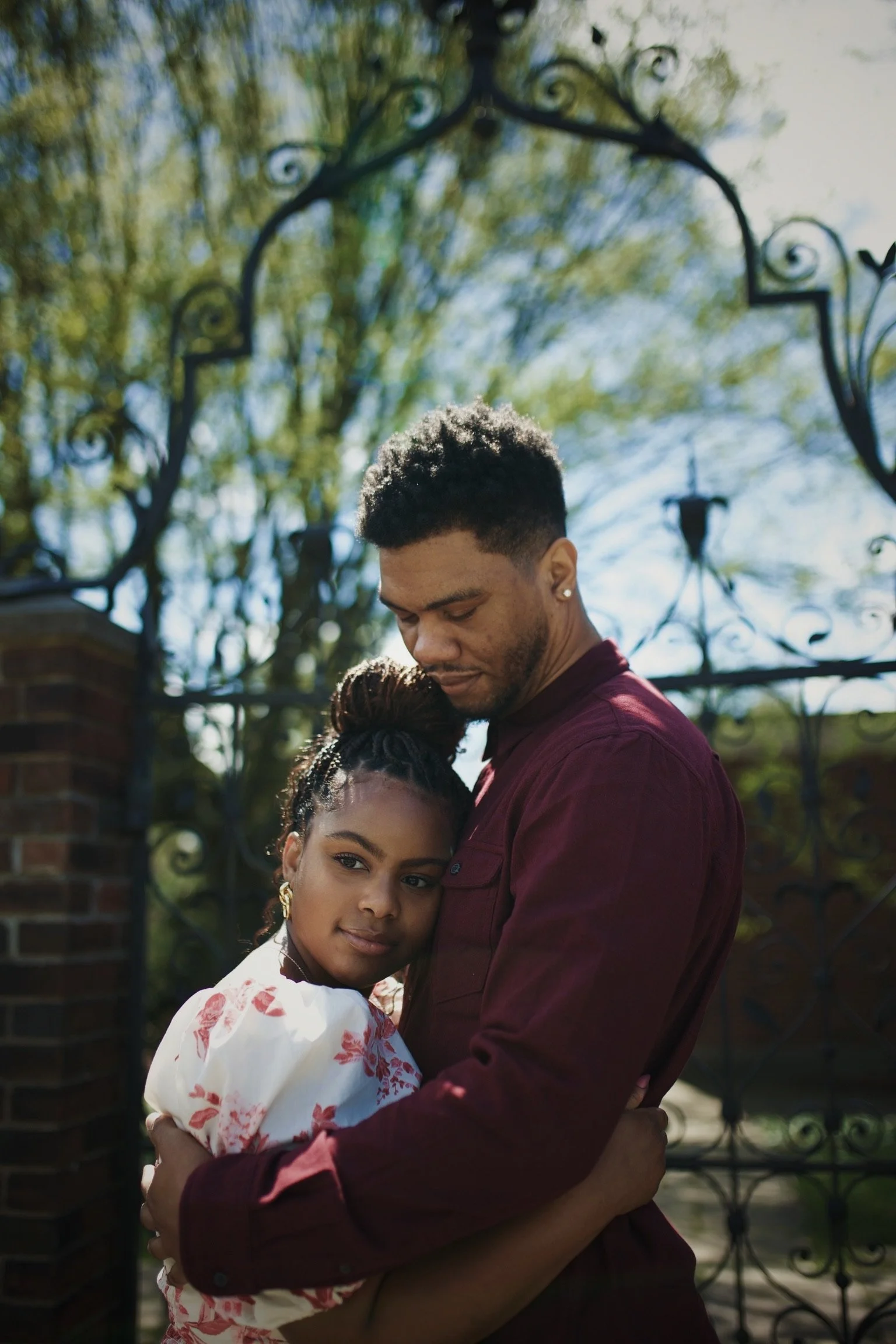 A young couple hugging outdoors, with a wrought iron gate and trees in the background.