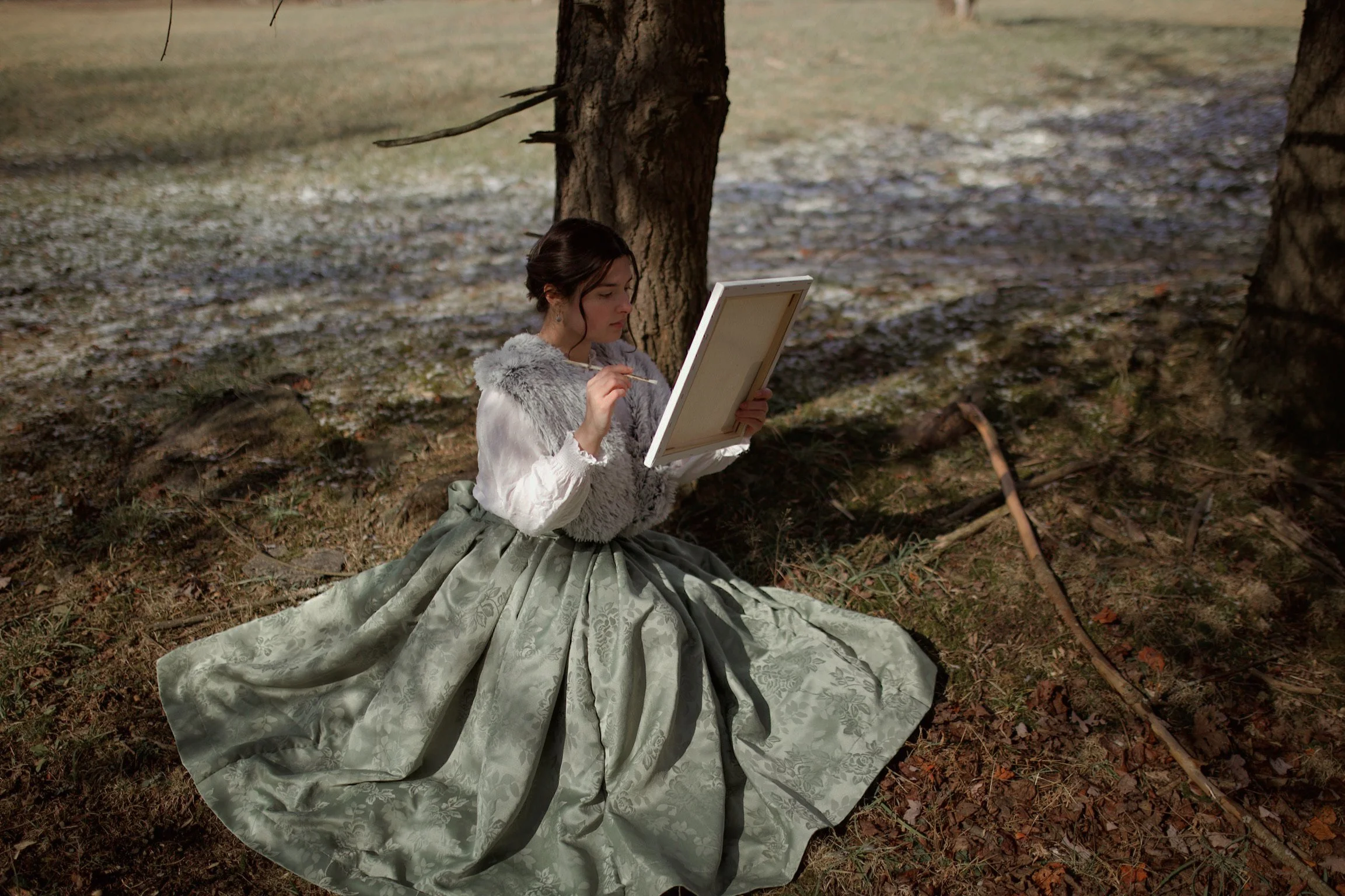A woman dressed in vintage clothing, with a full skirt and fur-lined top, sits on the ground under a tree in a forest, painting on a canvas.
