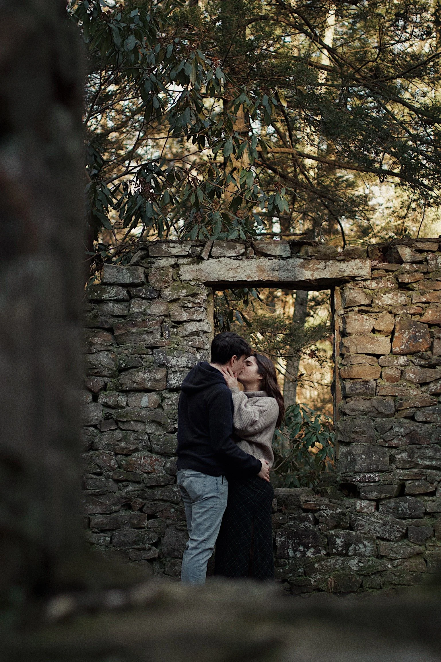 A young couple sharing a kiss and embracing in front of a rustic stone wall with a window opening, surrounded by trees and nature.