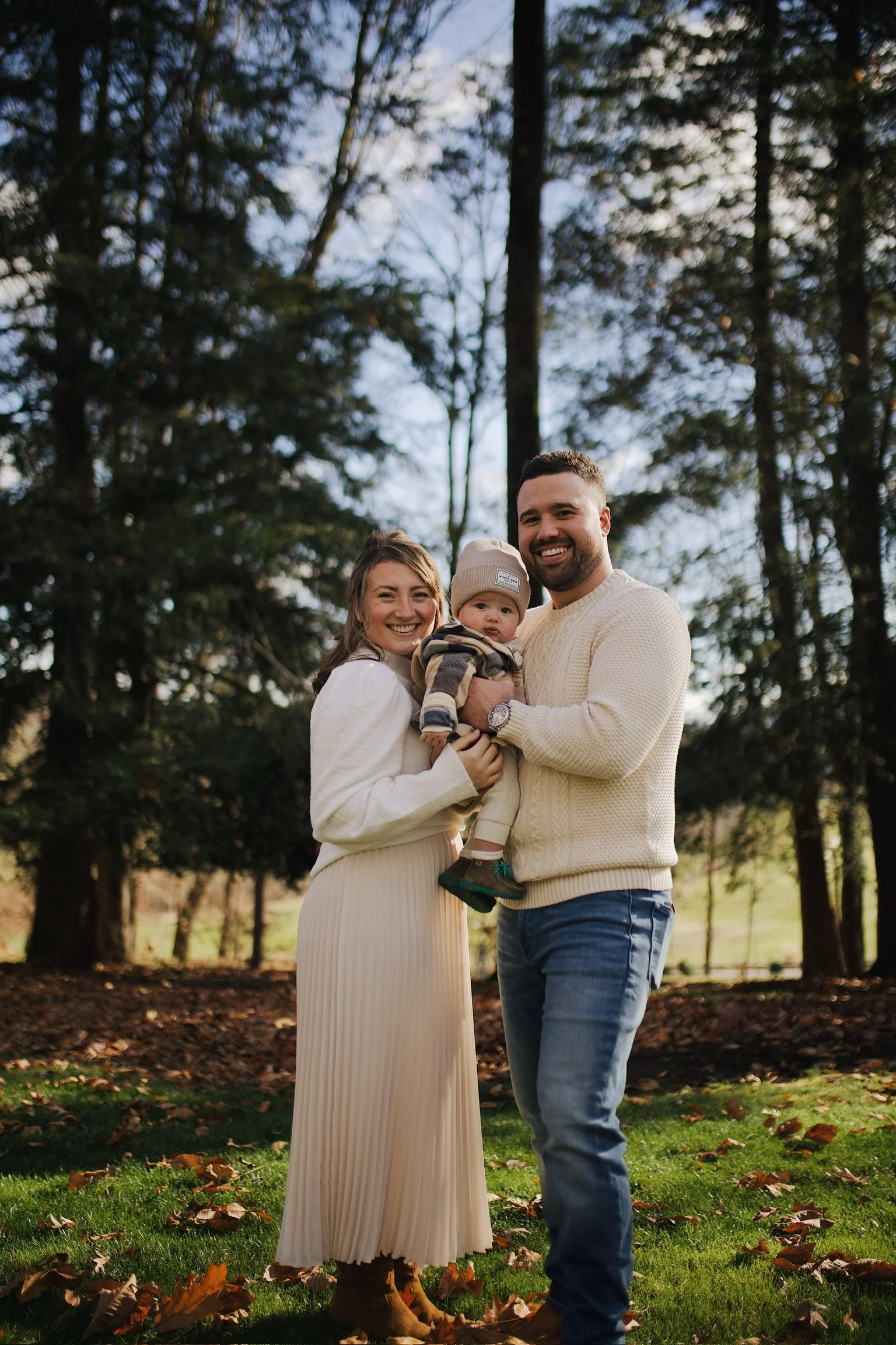A happy family of three outdoors in a wooded area during fall, with leaves on the ground, smiling and holding a young child.