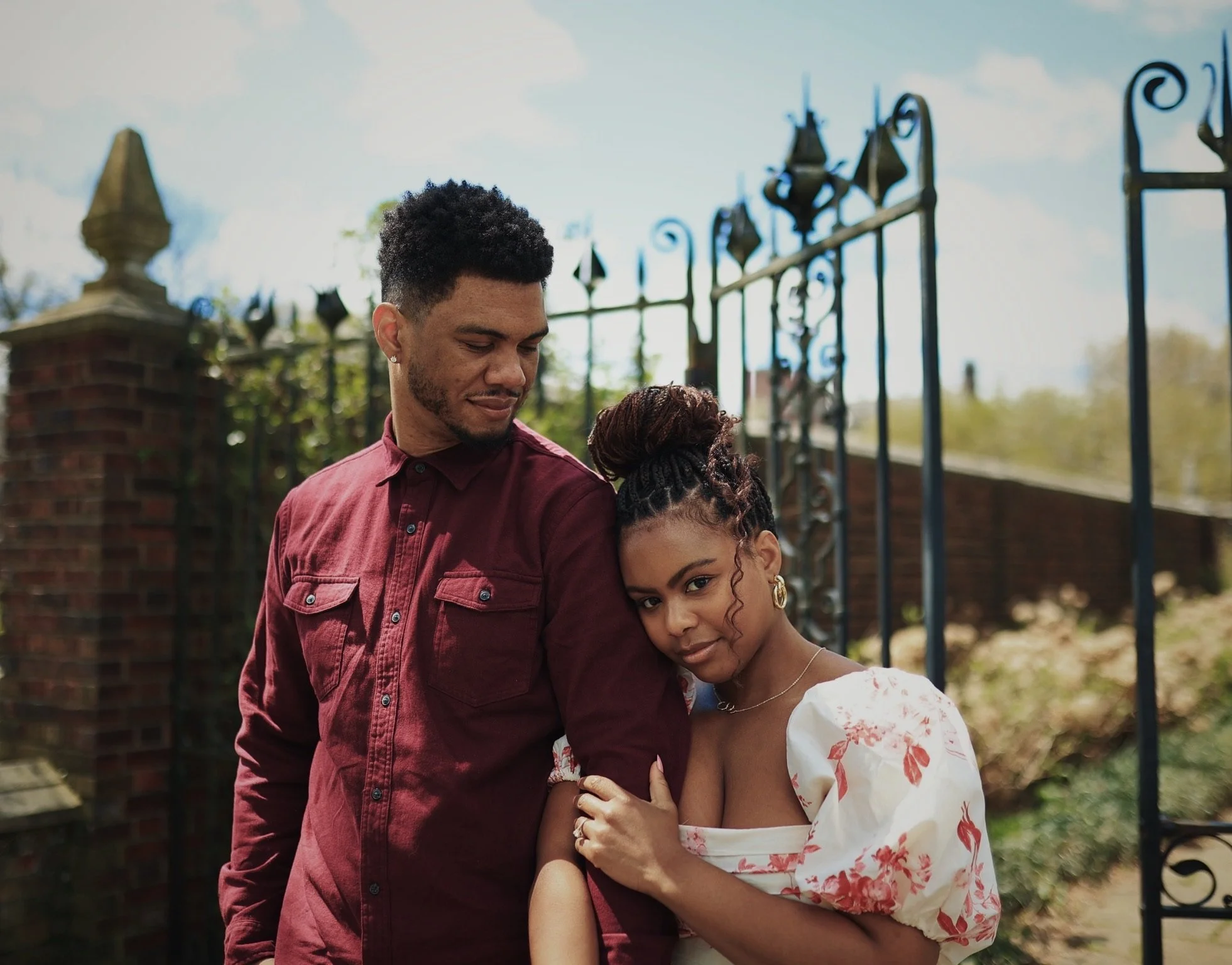 A young man with short curly hair and a young woman with braided hair and earrings are standing close together outdoors, with the woman leaning her head against the man's shoulder near a black iron gate and brick wall.
