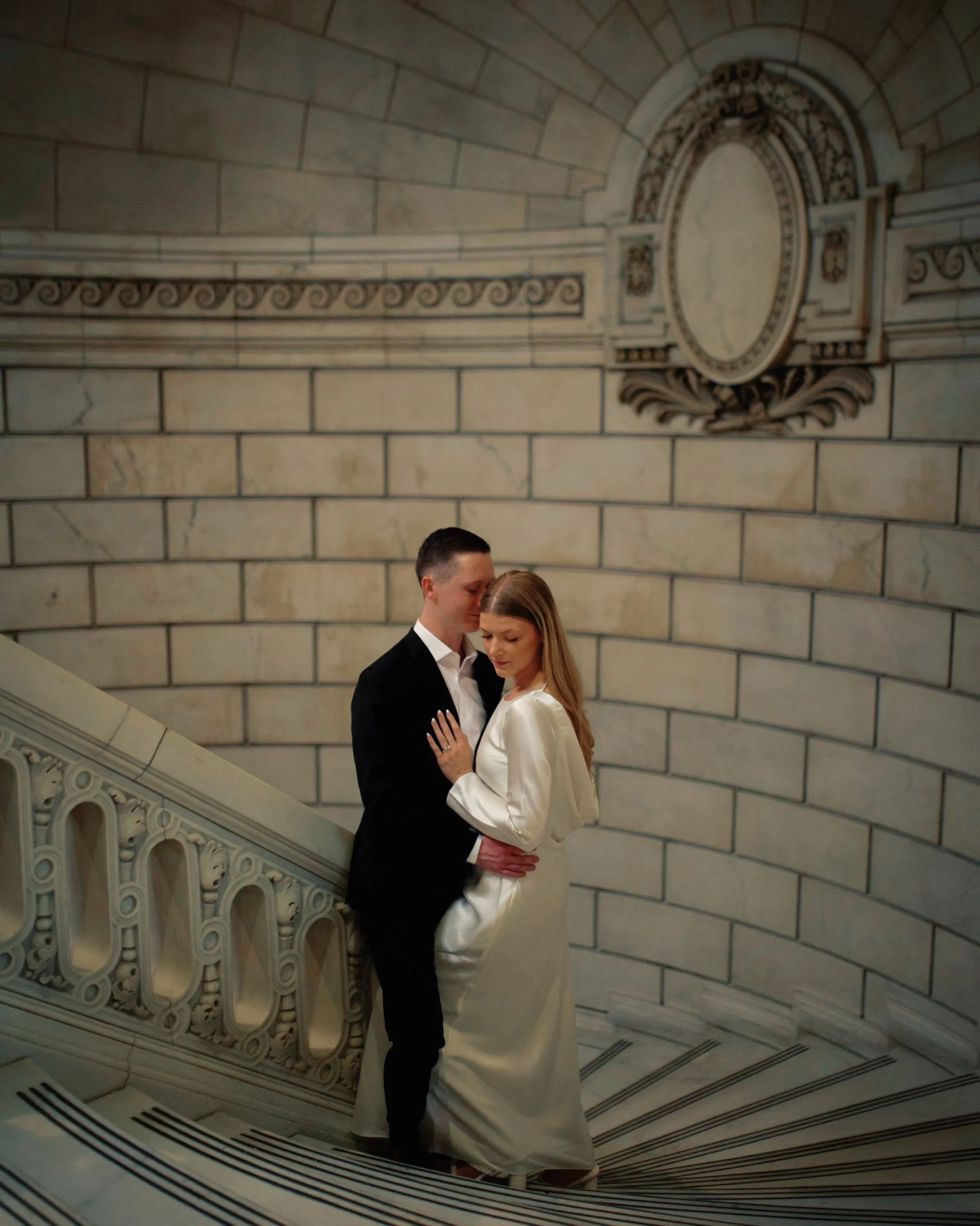 A man in a black suit and a woman in a white satin dress embrace on a curved staircase in a marble building with ornate wall decorations.