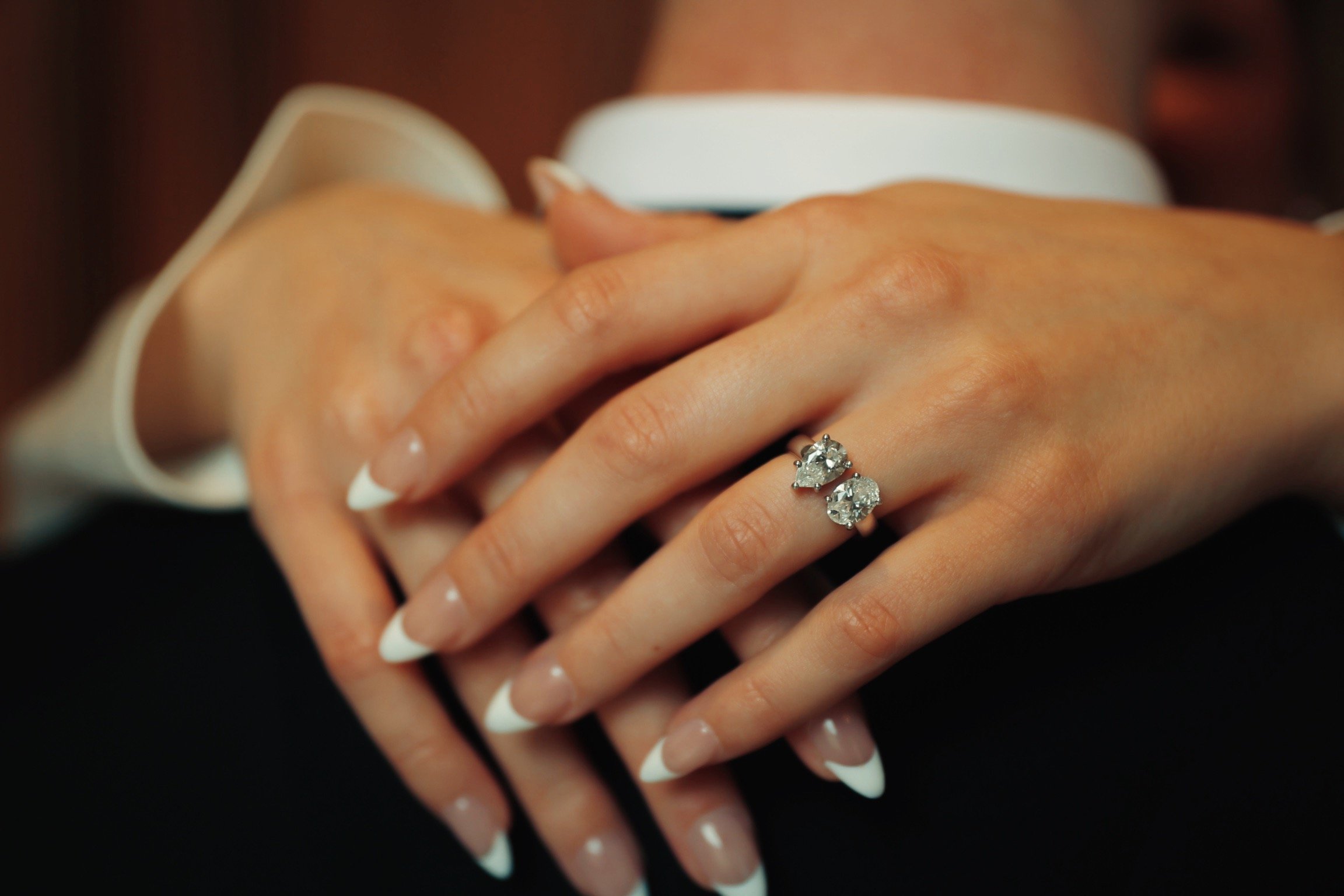 Close-up of hands with a diamond ring on the finger, showcasing a white long-sleeve shirt, blurred background.