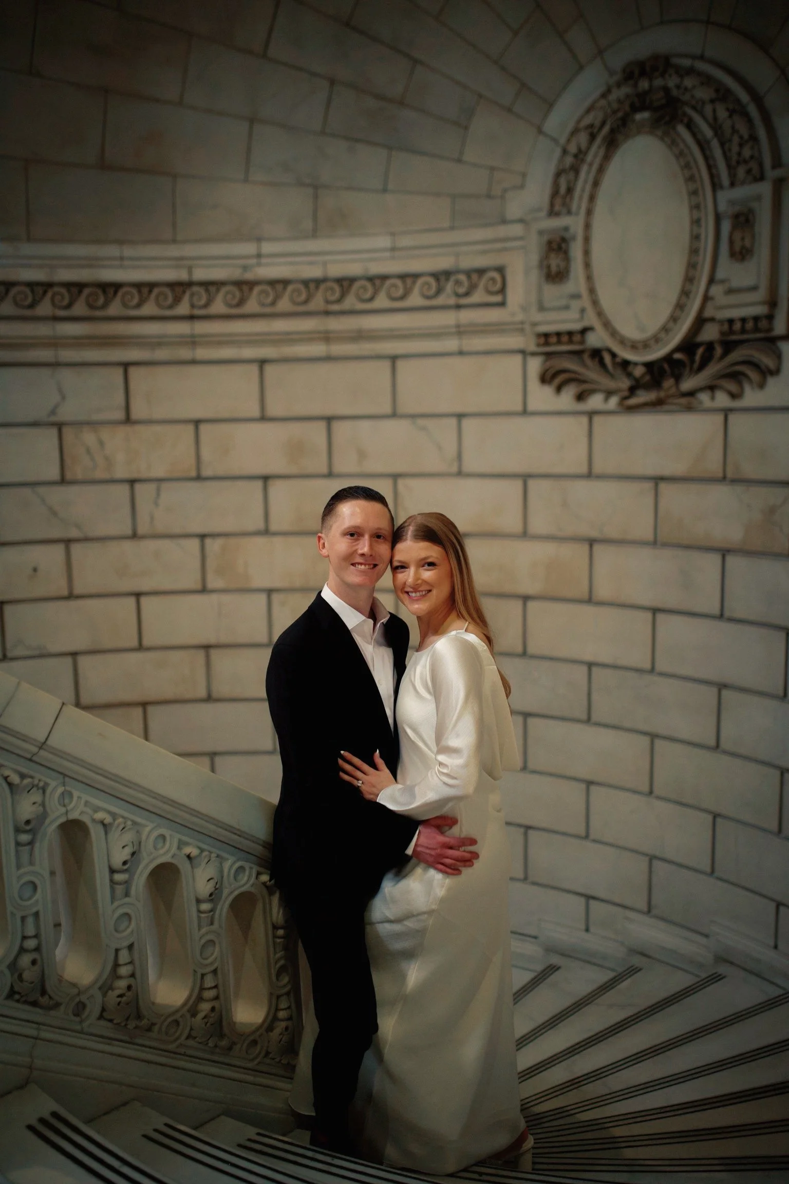 A smiling couple dressed in formal attire, standing on a decorative marble staircase inside a historic building with ornate wall and ceiling details.