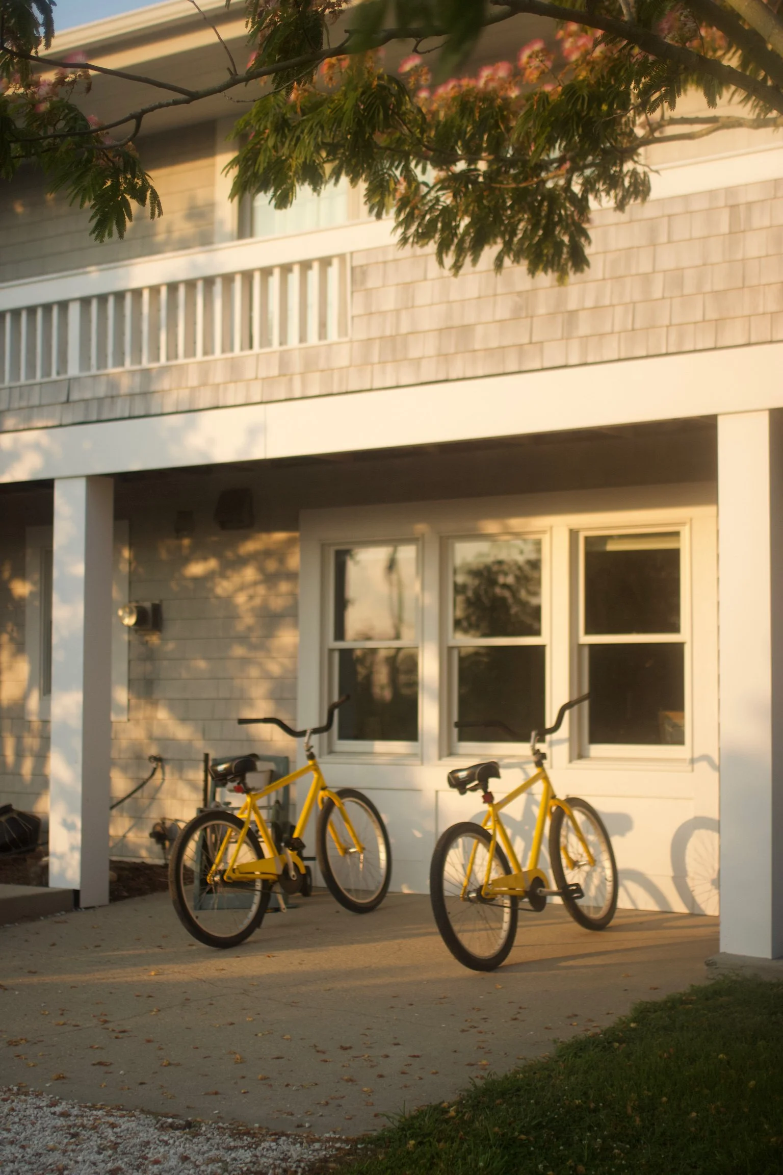 Two yellow bicycles parked on a concrete porch in front of a white house with three large windows, during sunset.