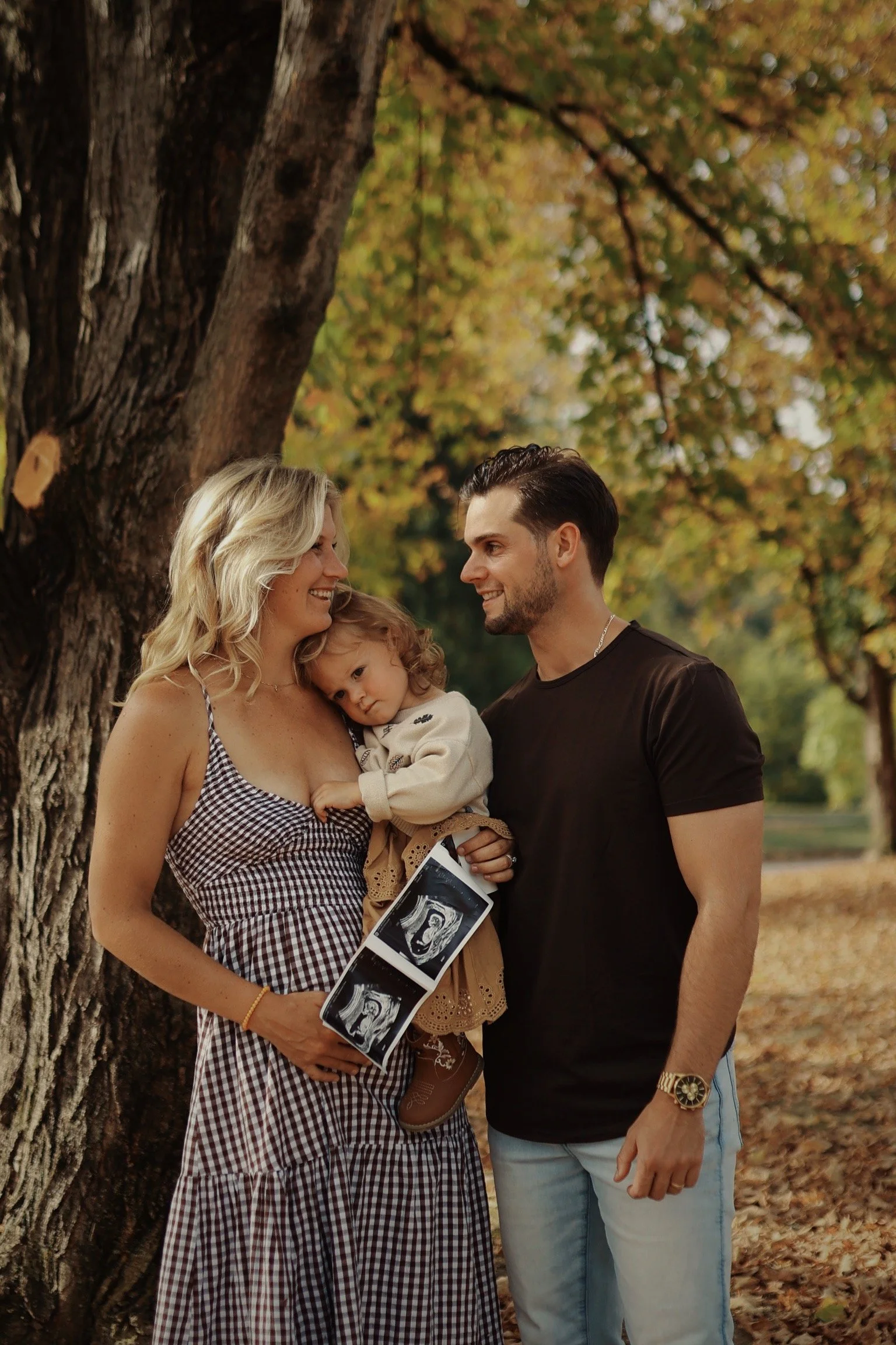 A family of three stands outdoors in a park during autumn, with colorful leaves on trees and the ground. The mother holds ultrasound photos, the daughter rests her head on the mother's chest, and the father looks at the mother with a smile.