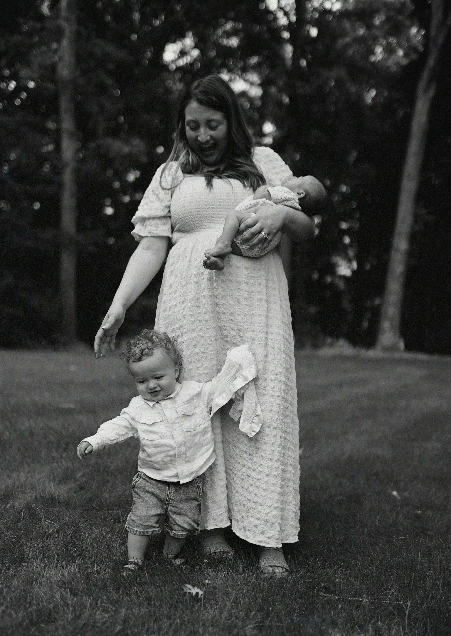 A woman in a long dress holds a sleeping baby in her arms while a young child, with curly hair, holding her hand, is standing on the grass in an outdoor setting with trees in the background, black and white photograph.