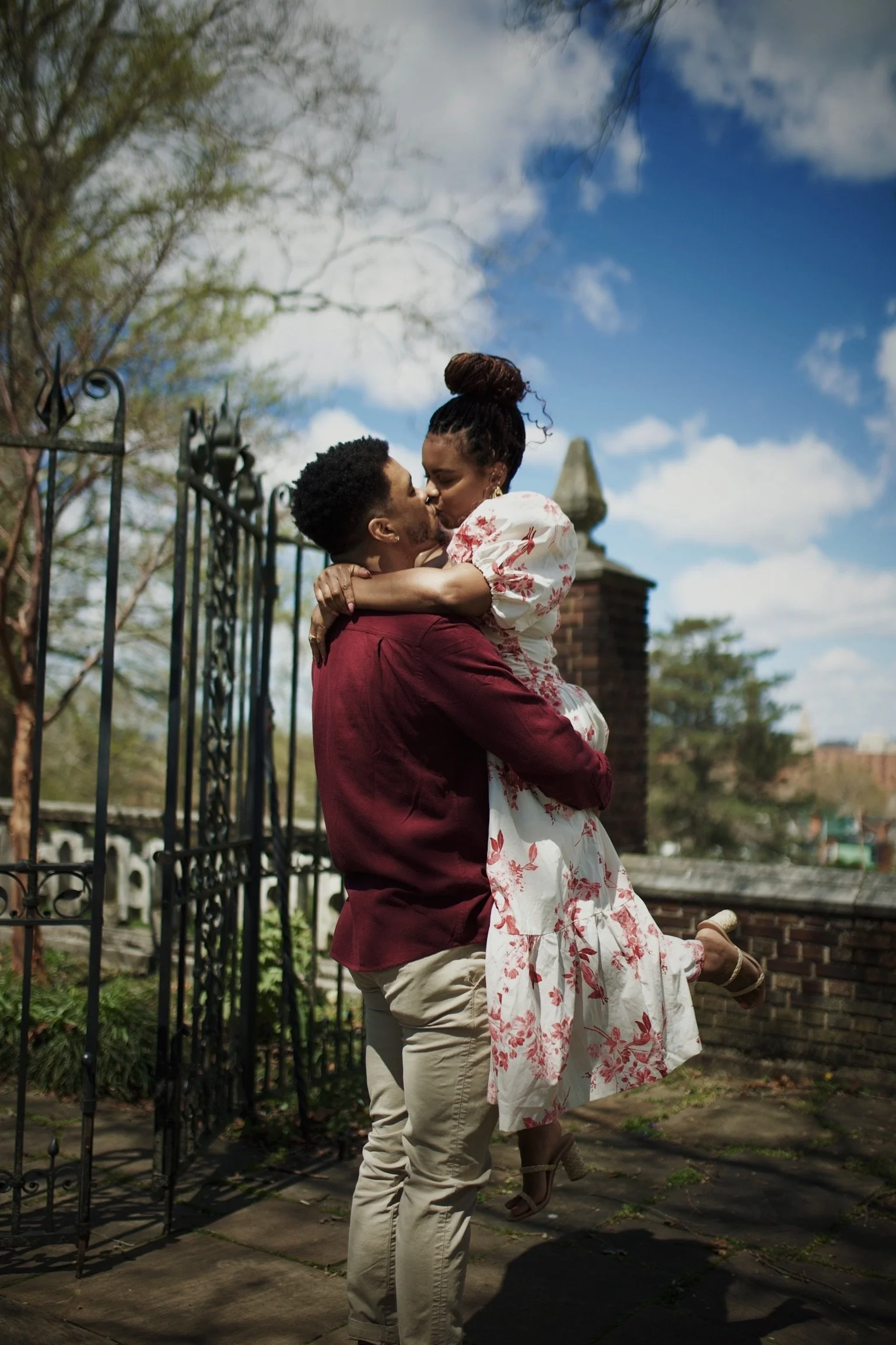 A couple sharing a kiss outdoors, with the woman being lifted in the man's arms against a backdrop of a brick wall, trees, and a partly cloudy sky.