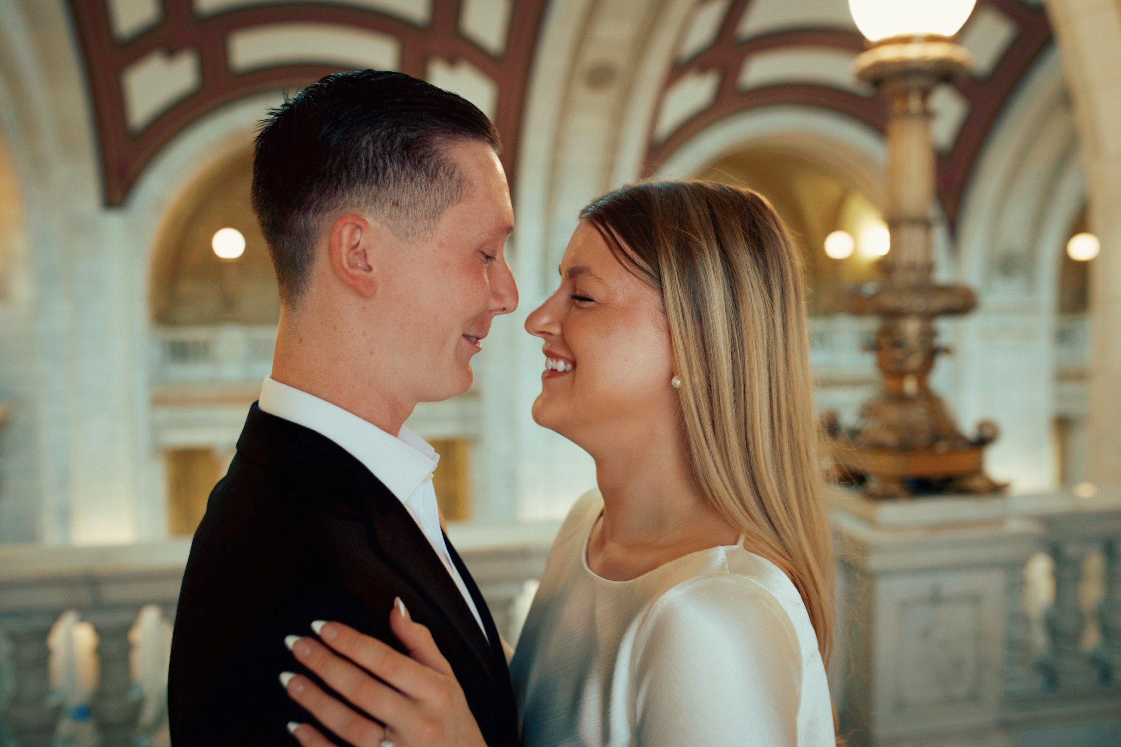 A couple smiling and about to kiss in an elegant, historic building with arched ceilings and a large central lamp.