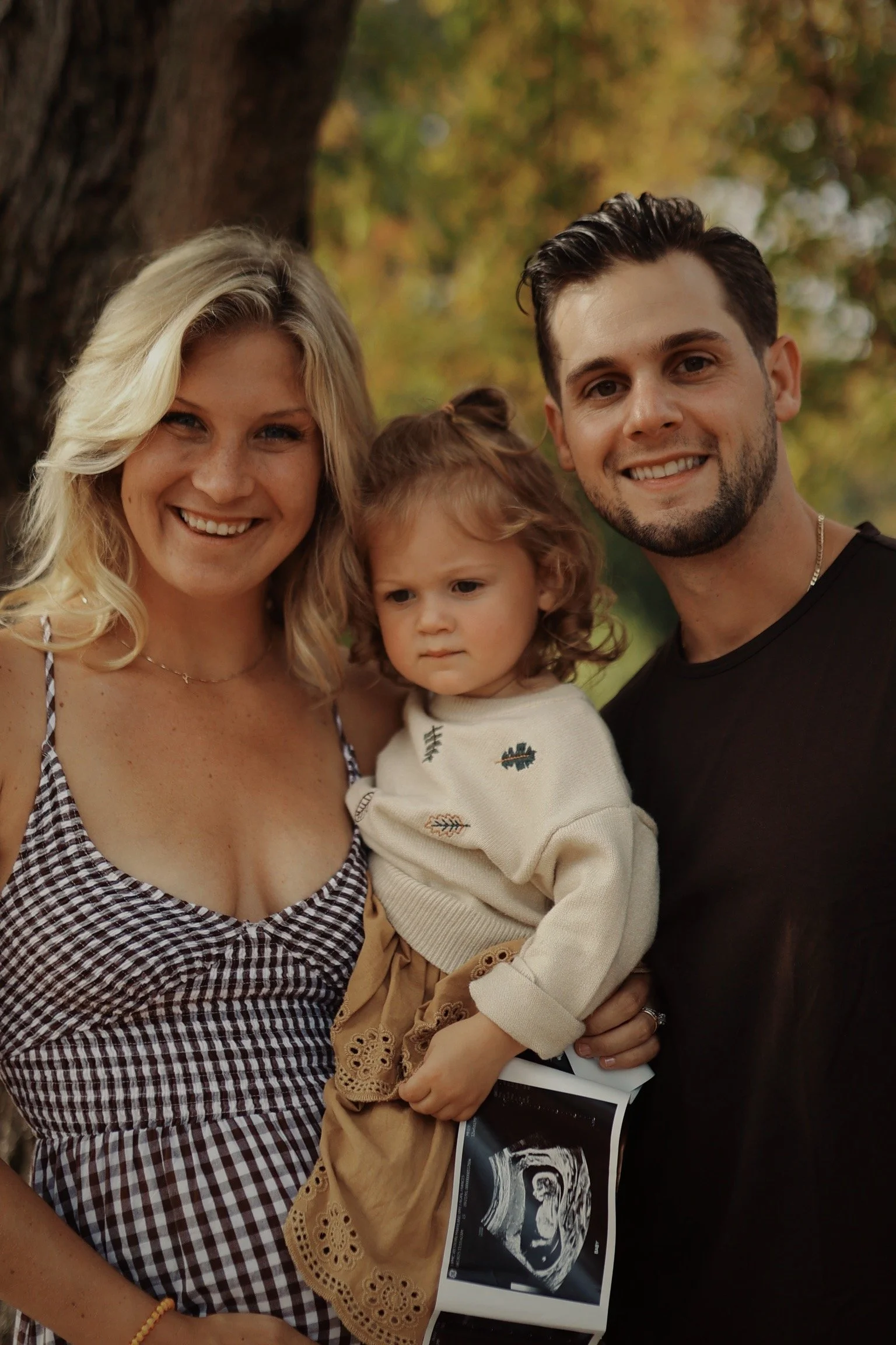 A happy family of three, a woman, a man, and a young girl, posing outdoors near a tree during autumn. The woman has blonde hair, smiling, wearing a checked dress. The man has dark hair and a beard, smiling, wearing a black shirt. The girl has curly h