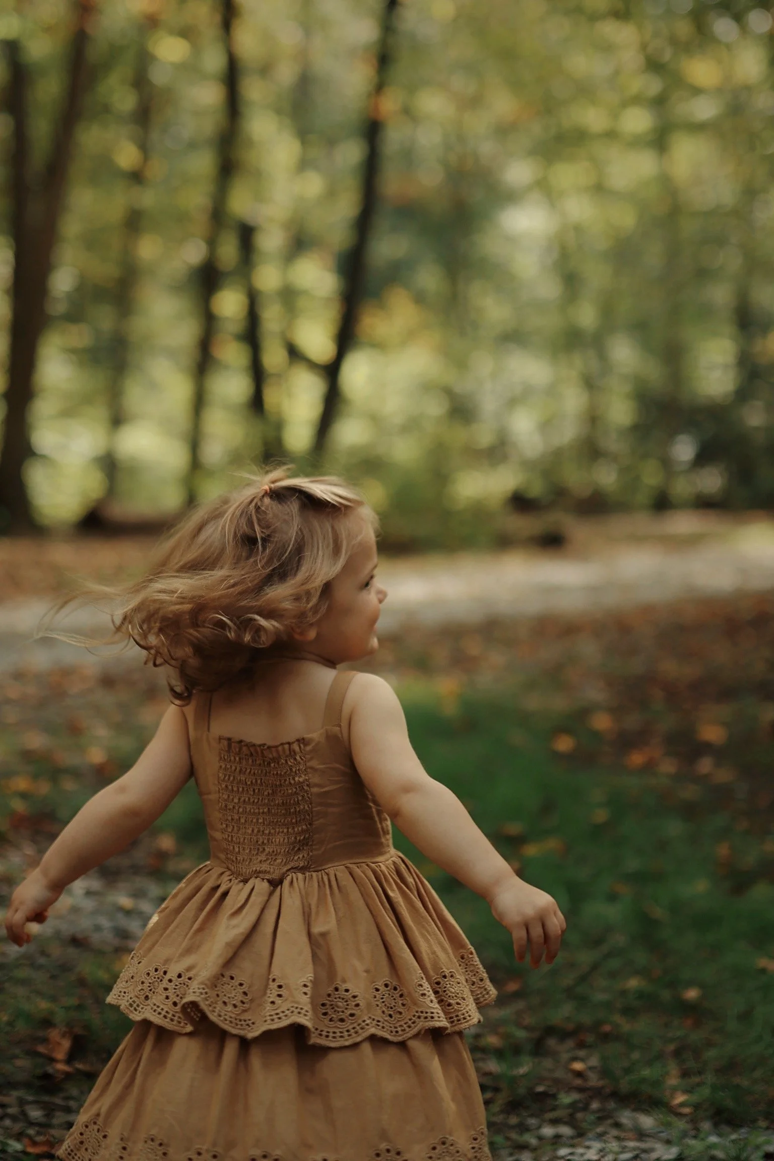 A young girl with curly blonde hair playing outdoors in a wooded area, wearing a tan dress with lace details.