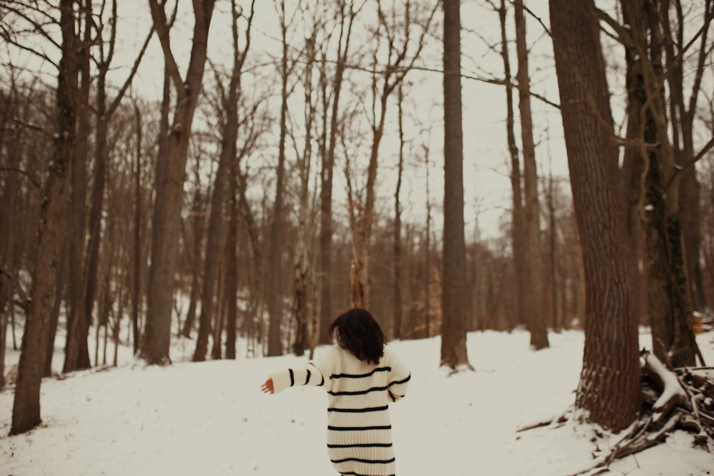 A person with dark hair wearing a cream and black striped sweater walking in a snow-covered forest with tall, leafless trees.