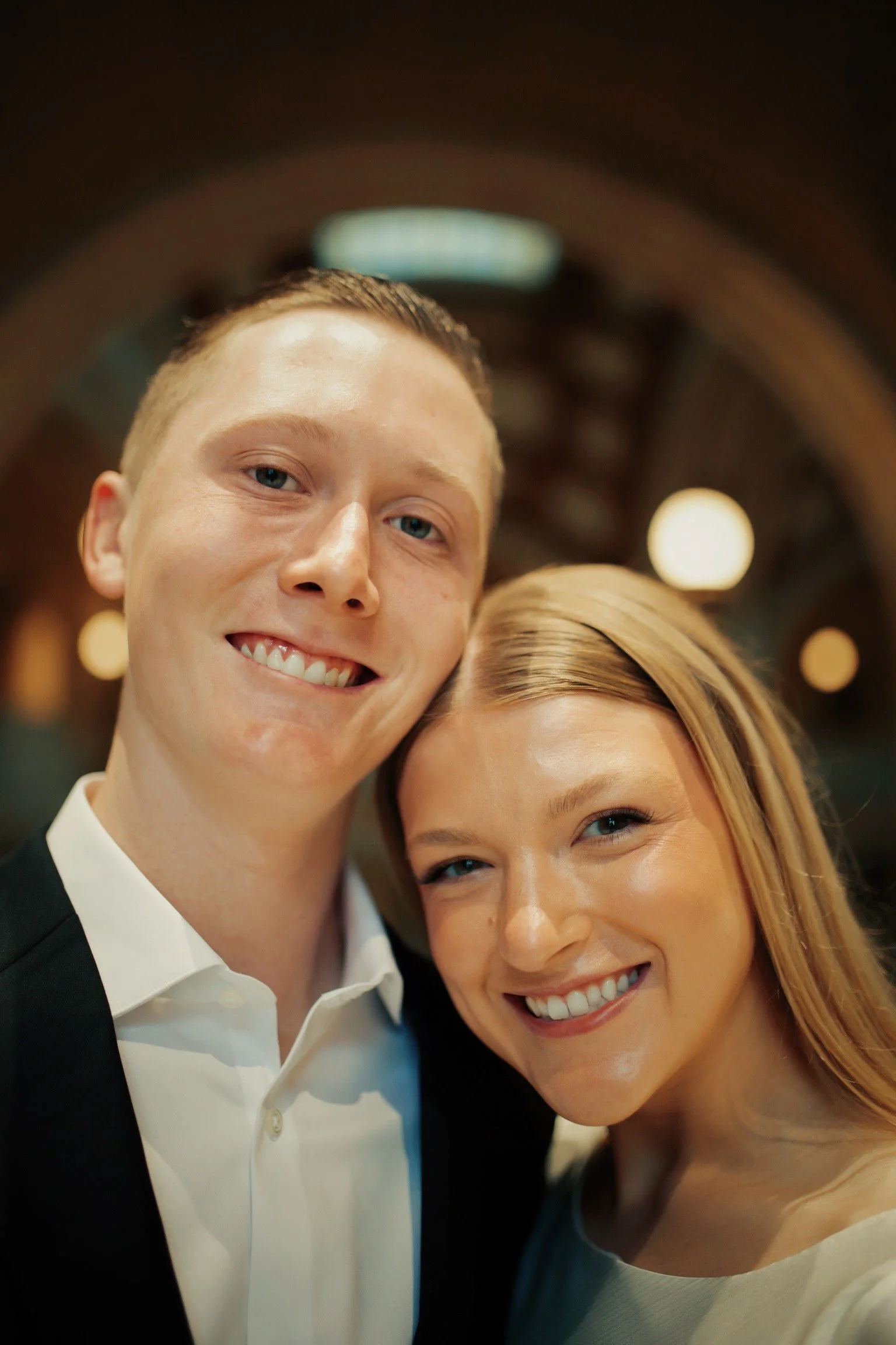 Close-up of smiling young man and woman taking a selfie together indoors, with blurred warm lights in the background.